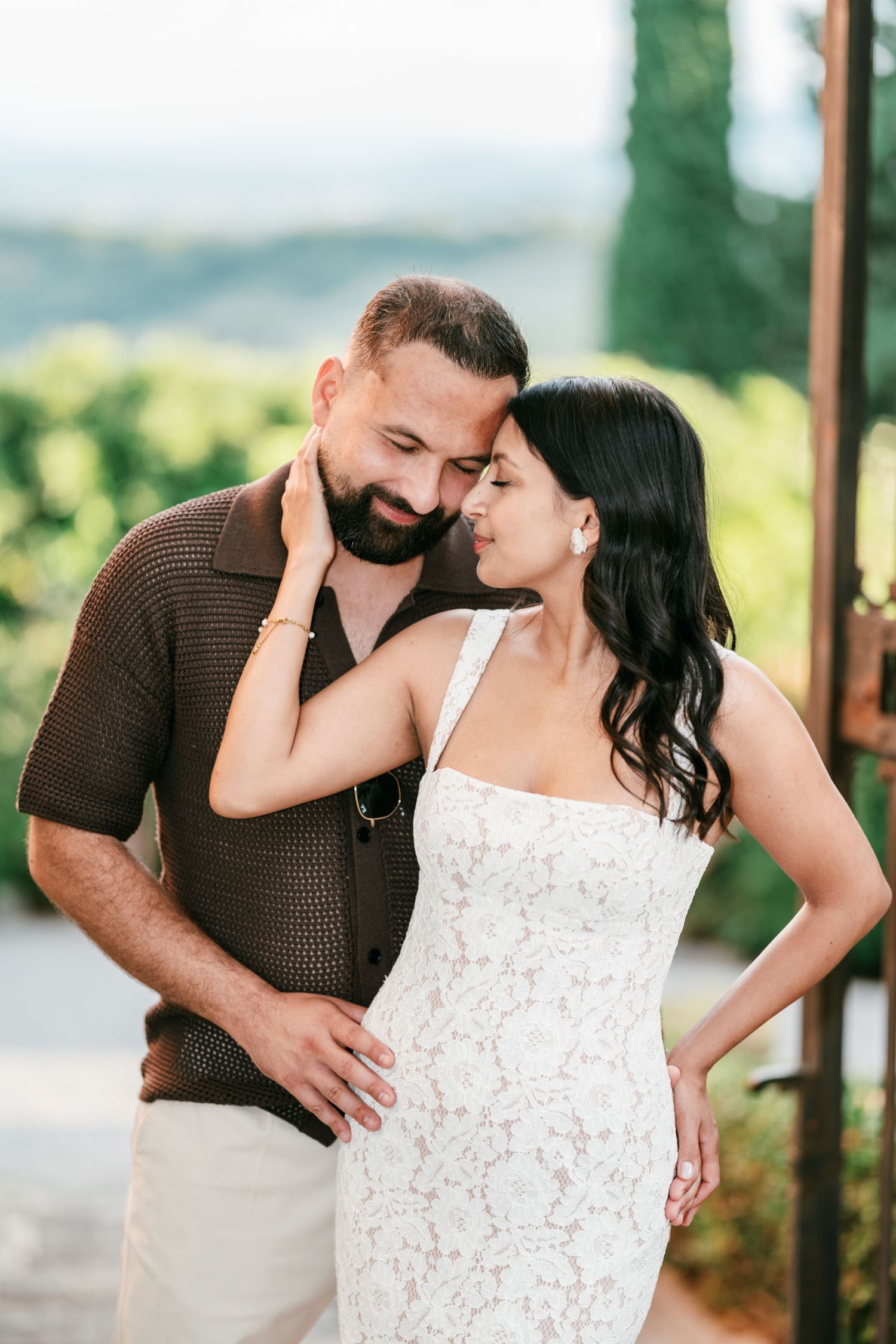 A bearded man in a brown knit shirt rests his forehead against a woman in a white lace dress in a scenic hillside setting.