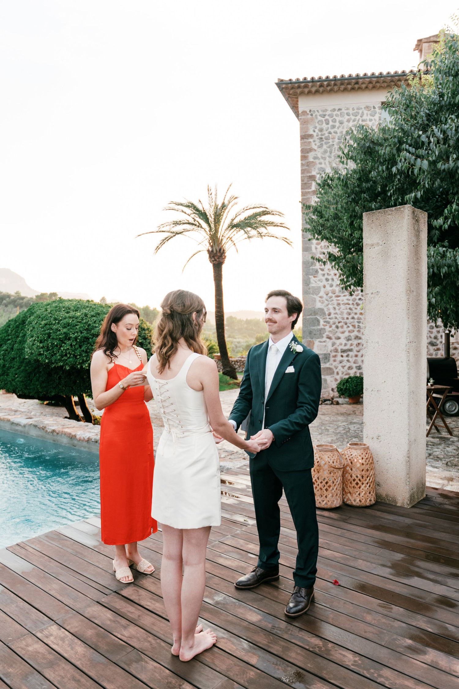 Couple holds hands during an outdoor wedding ceremony on a wooden deck by a pool as an officiant in a red dress smiles beside them.