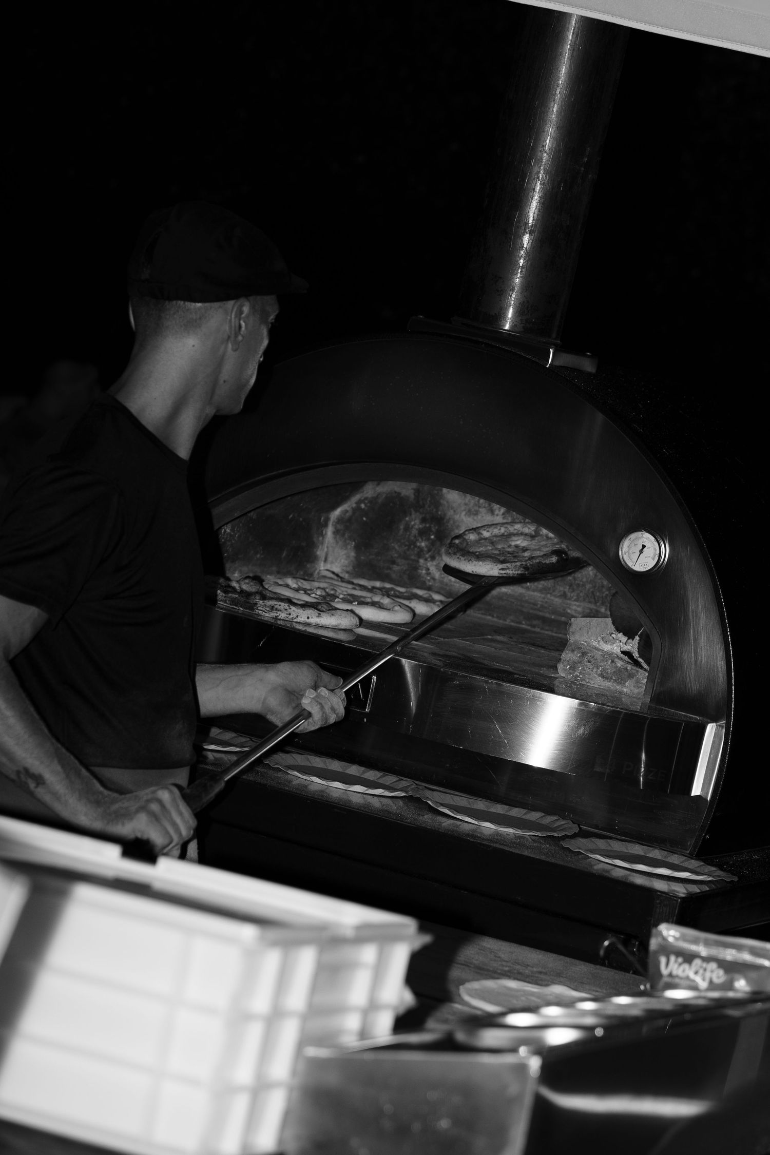 Black and white photo of a pizza maker using a long peel to tend pizzas inside a wood-fired oven with a chimney.