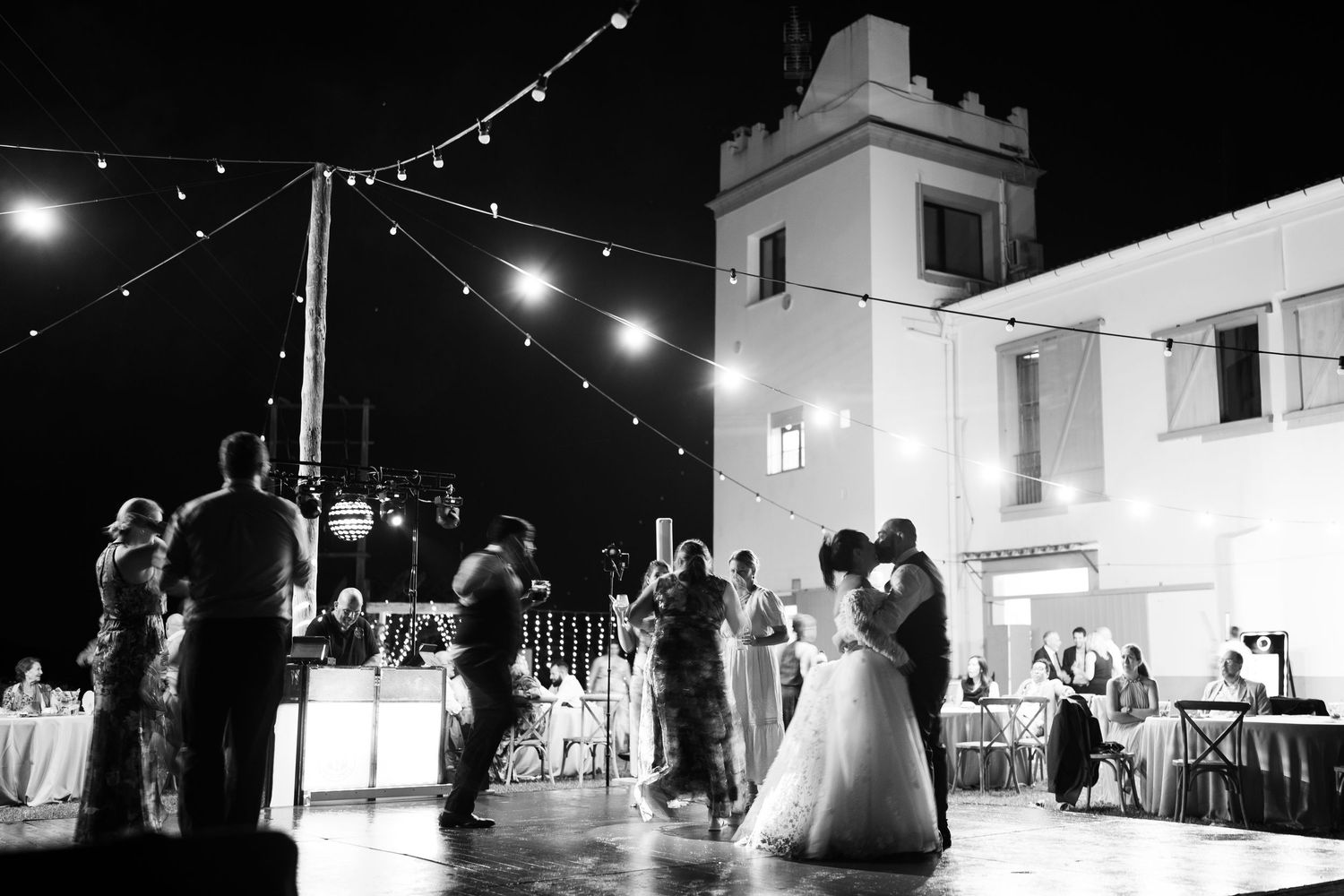 Black and white photo of a bride and groom sharing a first dance under string lights as guests watch outside a white tower building.