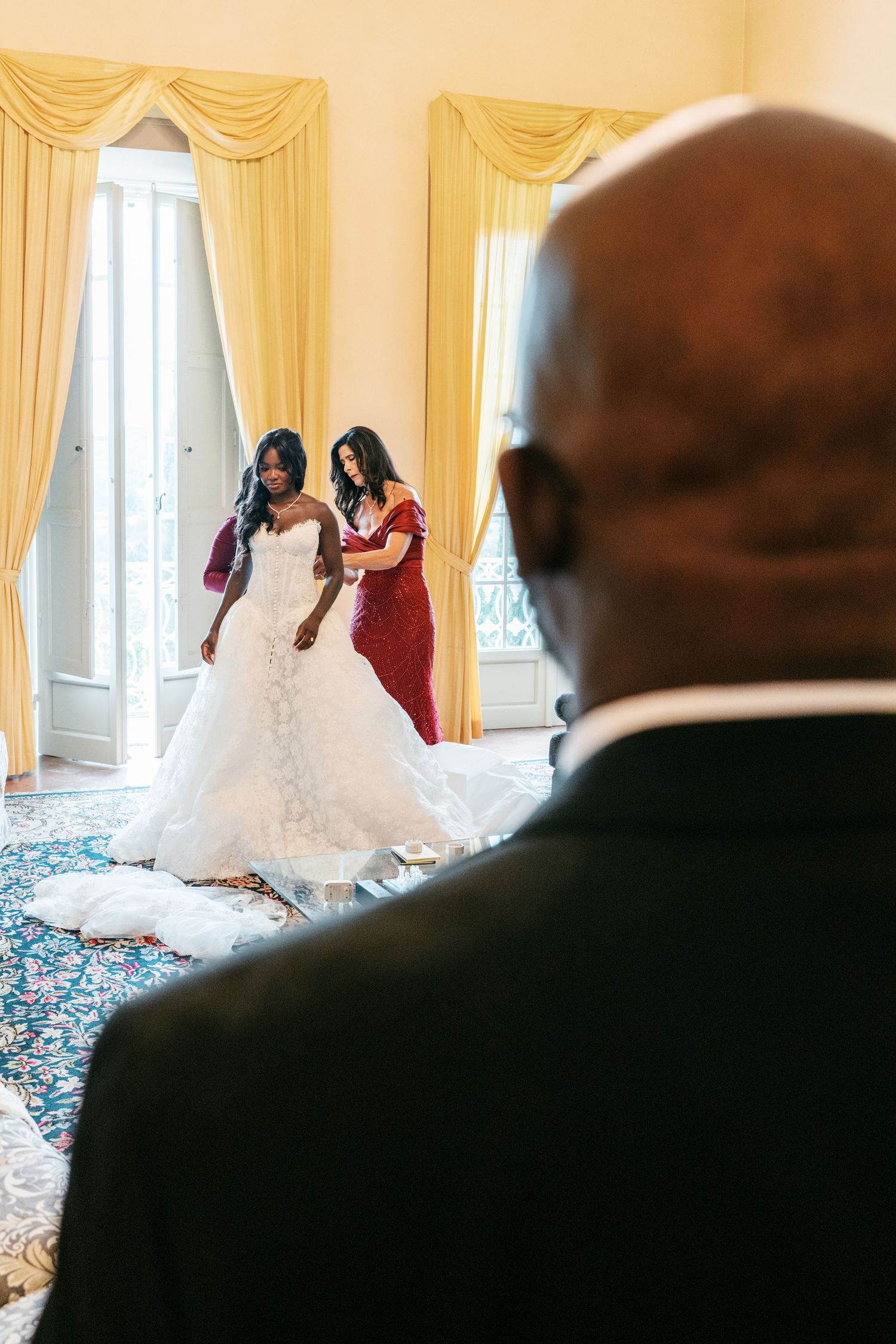 Bride in a white strapless wedding gown gets help from a bridesmaid in red as a man watches in a bright room with yellow drapes.