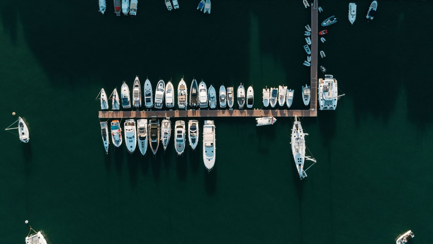 Aerial view of numerous small boats moored along a wooden pier in dark green harbor water.