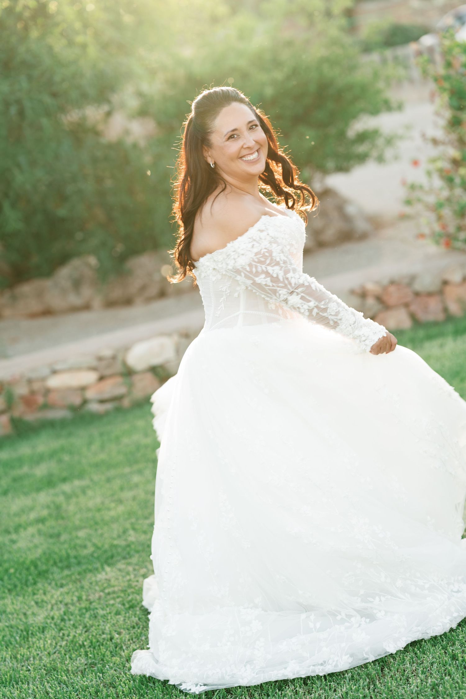 Smiling bride in a white lace off-the-shoulder wedding gown twirls on a sunlit green lawn beside a rustic stone wall.
