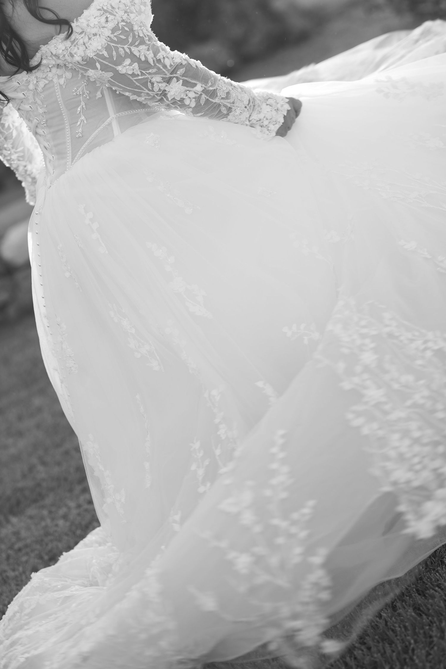 Black and white photo of a bride in a flowing tulle wedding gown with lace floral appliqués and long lace sleeves on grass.