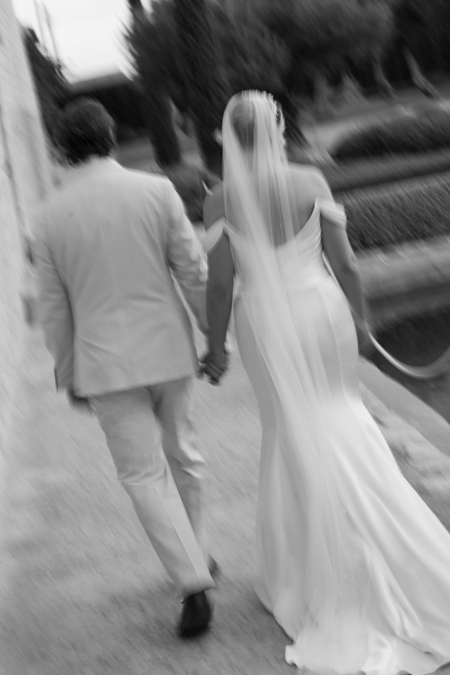 Black and white photo of a bride in a long veil and gown holding hands with a groom in a light suit as they walk away.