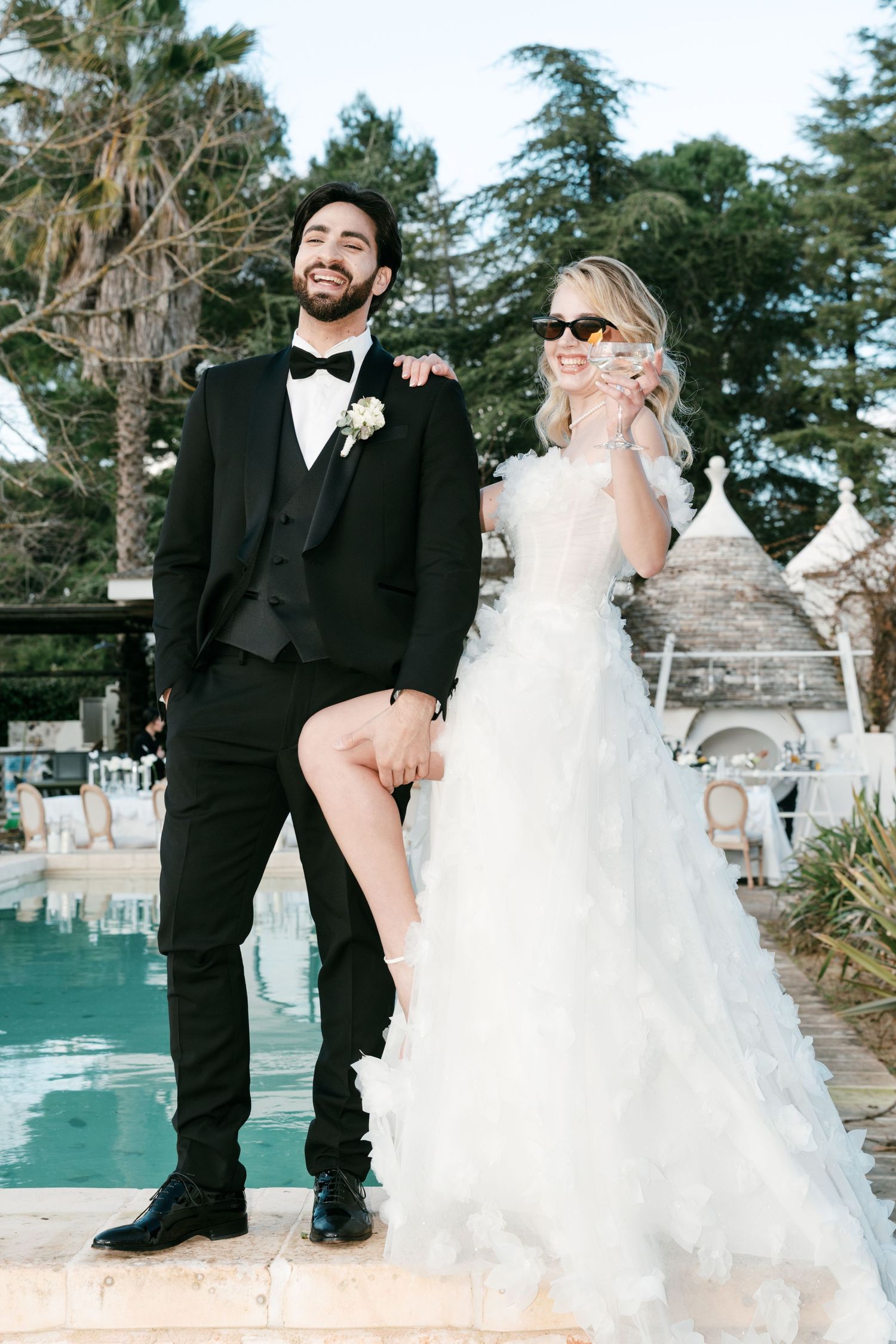 Bride in white ruffled gown and groom in black tuxedo pose joyfully by a pool at their outdoor wedding.