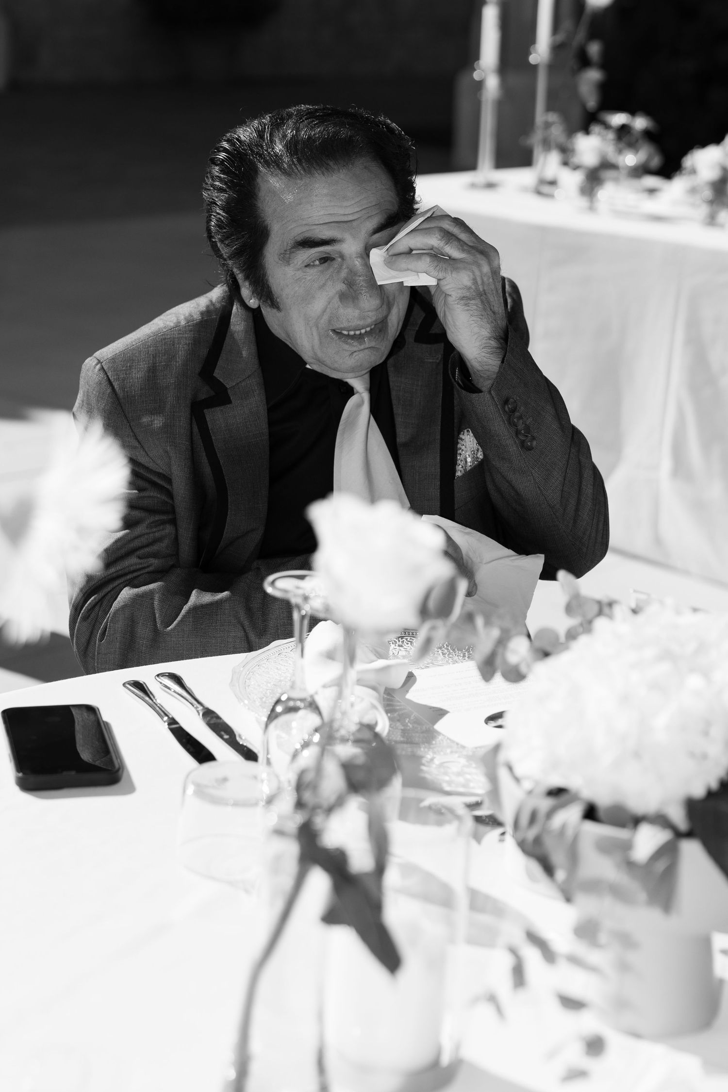Black and white photo of an emotional man wiping tears at a formal dinner table with flowers and candles in Mallorca.