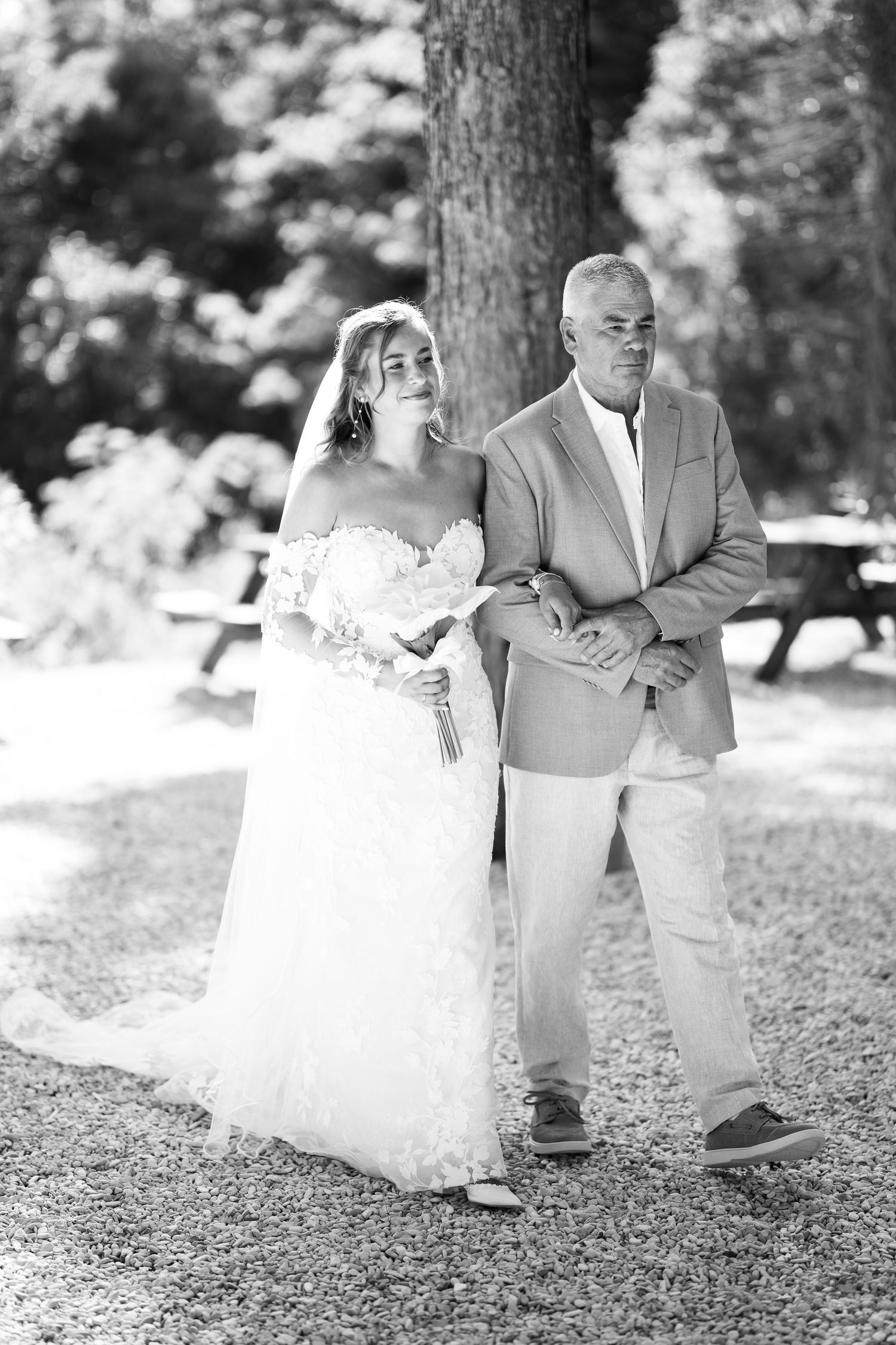 Black and white photo of a bride in a lace gown holding flowers, walking with an older man in a suit outdoors.
