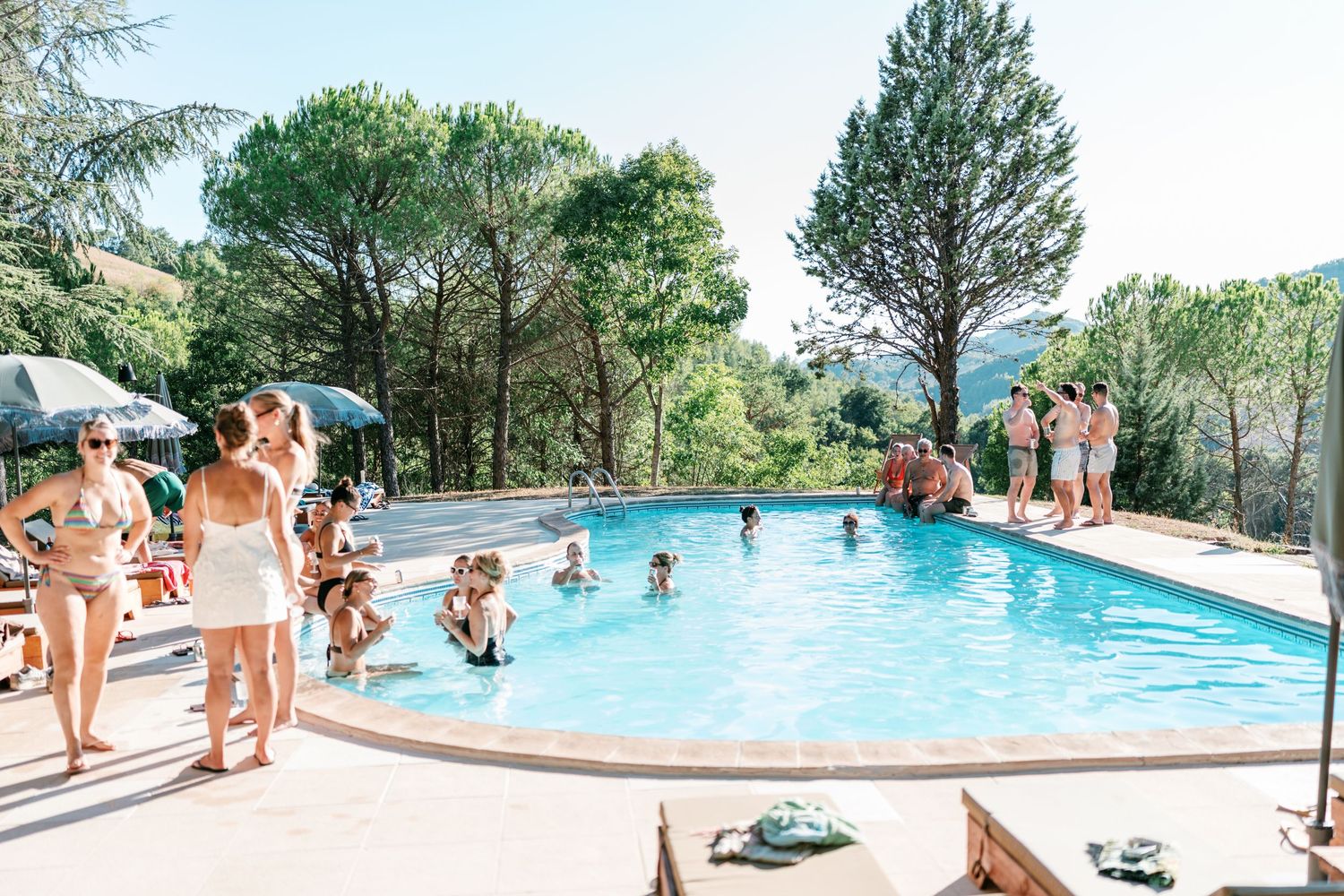 Outdoor swimming pool surrounded by trees with people swimming and relaxing on a sunny day at a resort.