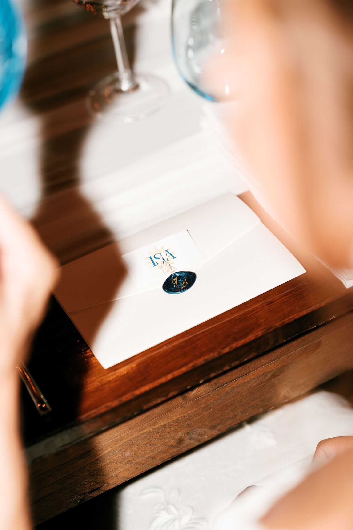 Close-up of an elegant wedding invitation with wax seal on a wooden box, with wine glasses in the background.