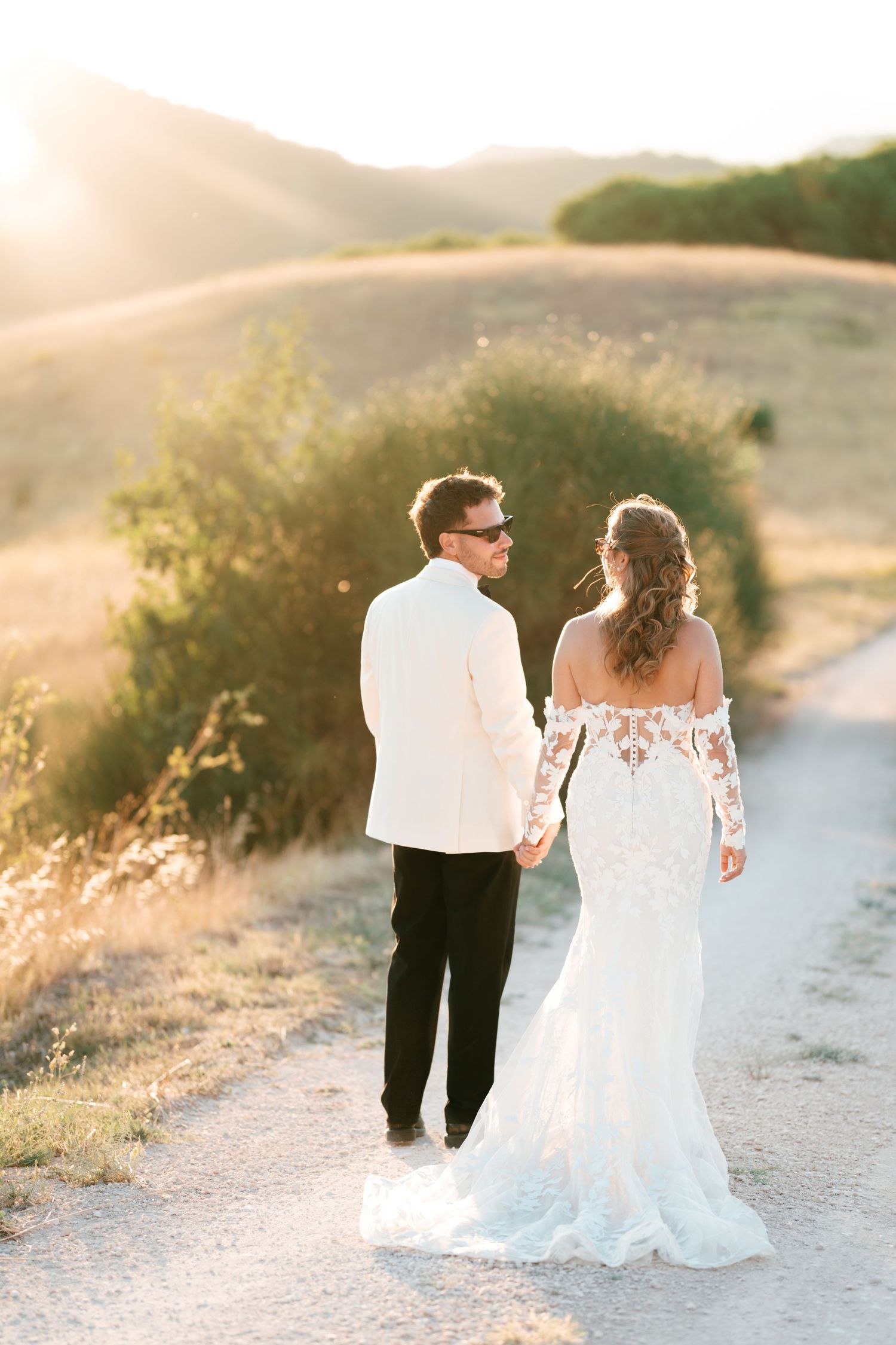 Bride and groom walking hand in hand on a sunlit path with golden hills at sunset.