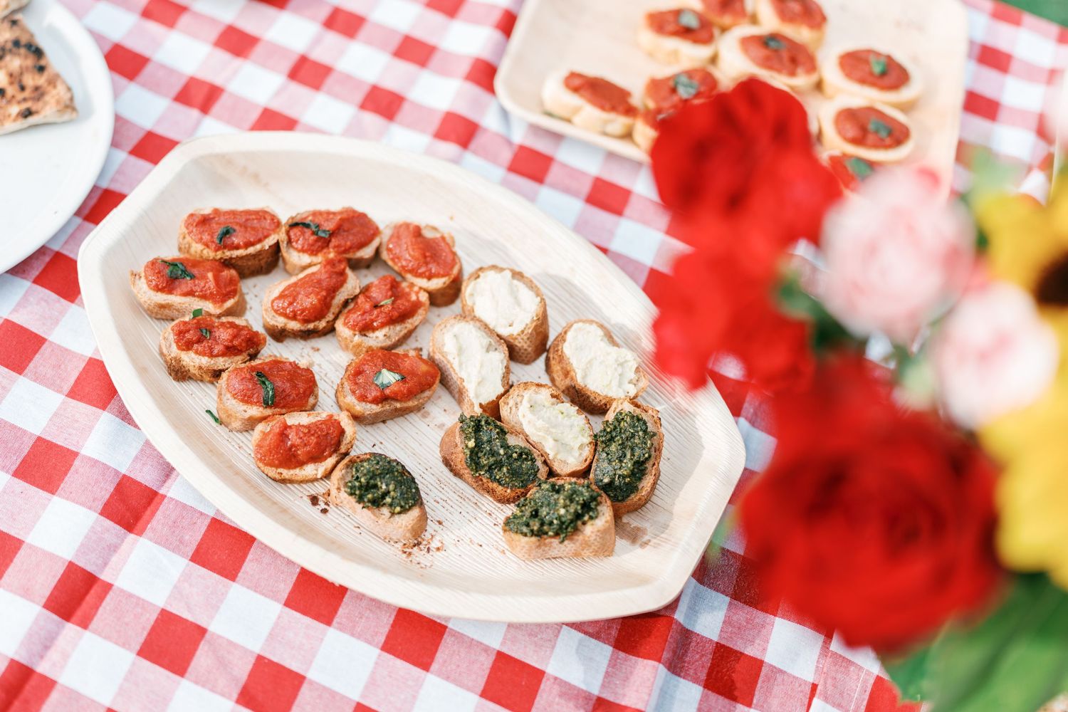Assorted bruschetta topped with tomato, pesto and cheese on a white platter over a red checkered tablecloth, with colorful flowers nearby.