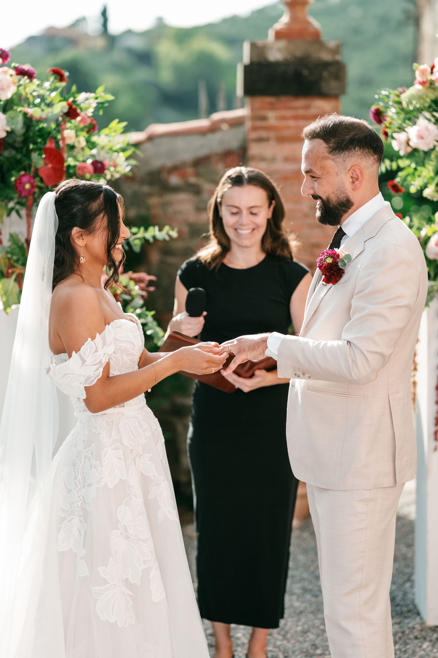 Bride in a lace off-shoulder gown and groom in a cream suit exchange rings during an outdoor ceremony led by a smiling officiant.