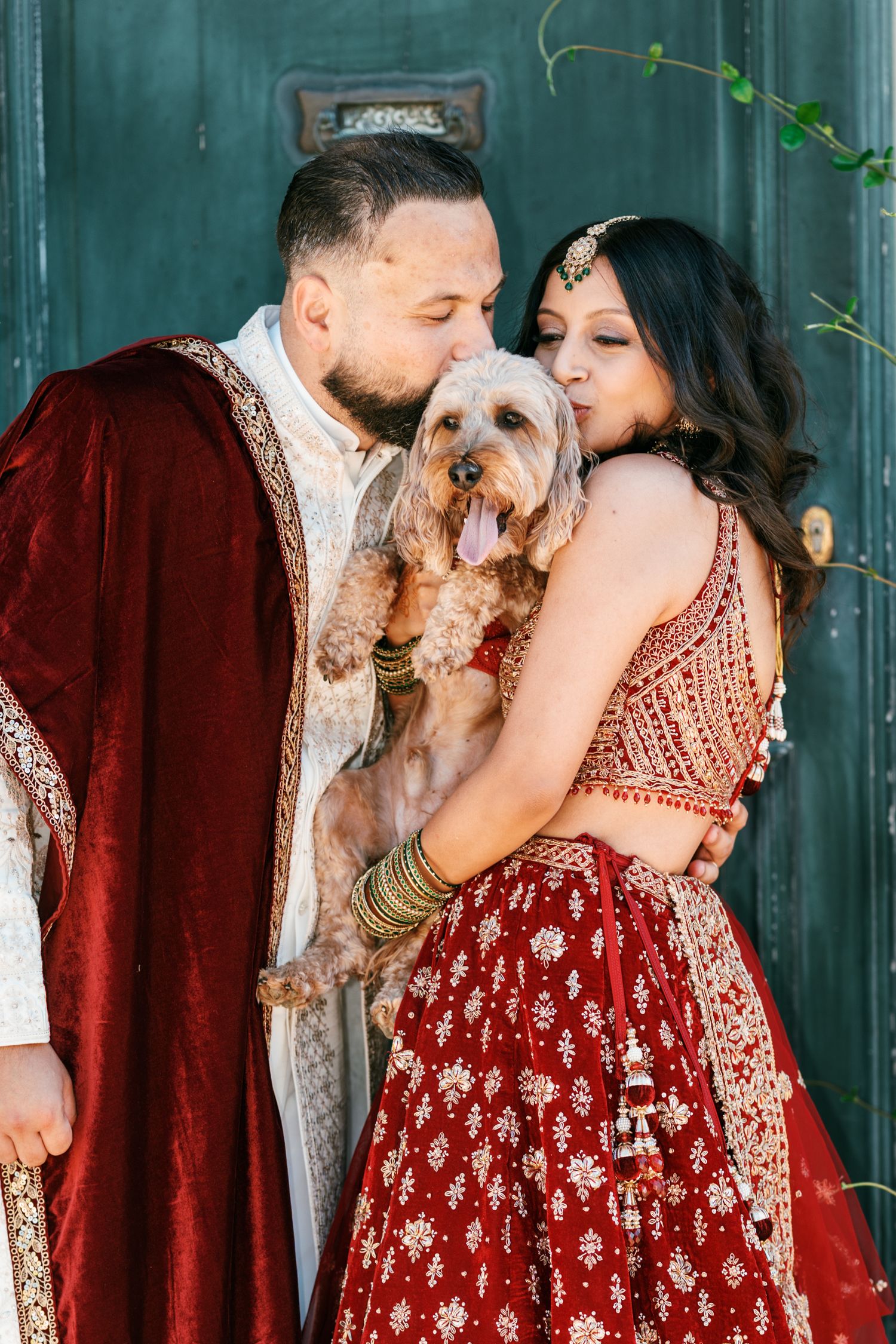 Bride and groom in red and gold Indian wedding attire kiss a fluffy dog held between them in front of a green door.