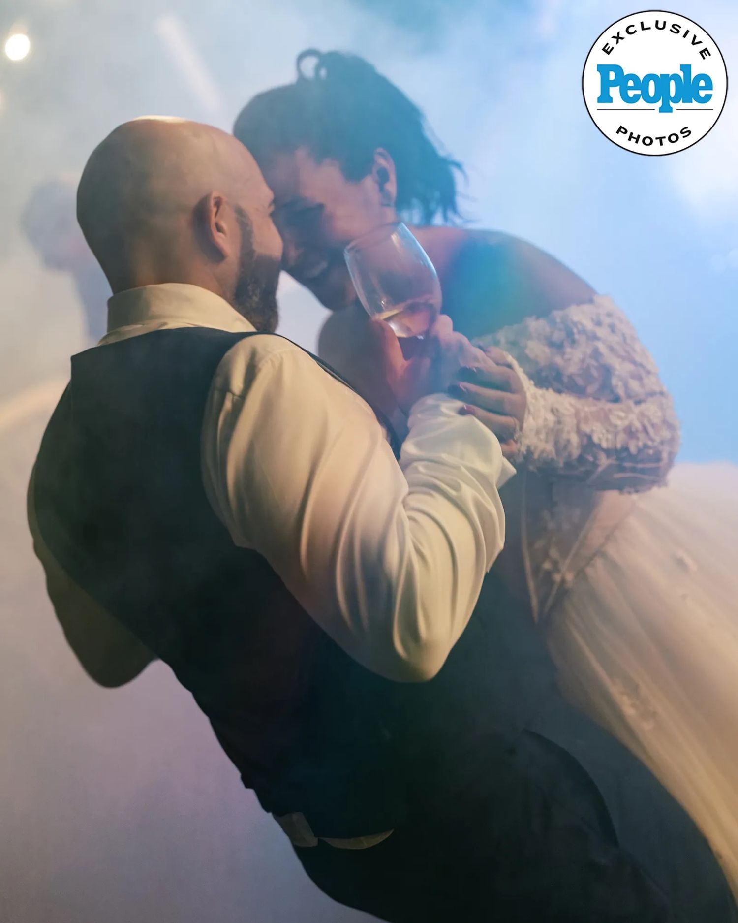 A bald, bearded groom in a vest and white shirt dances closely with a bride in a lace gown, holding a glass amid stage fog.