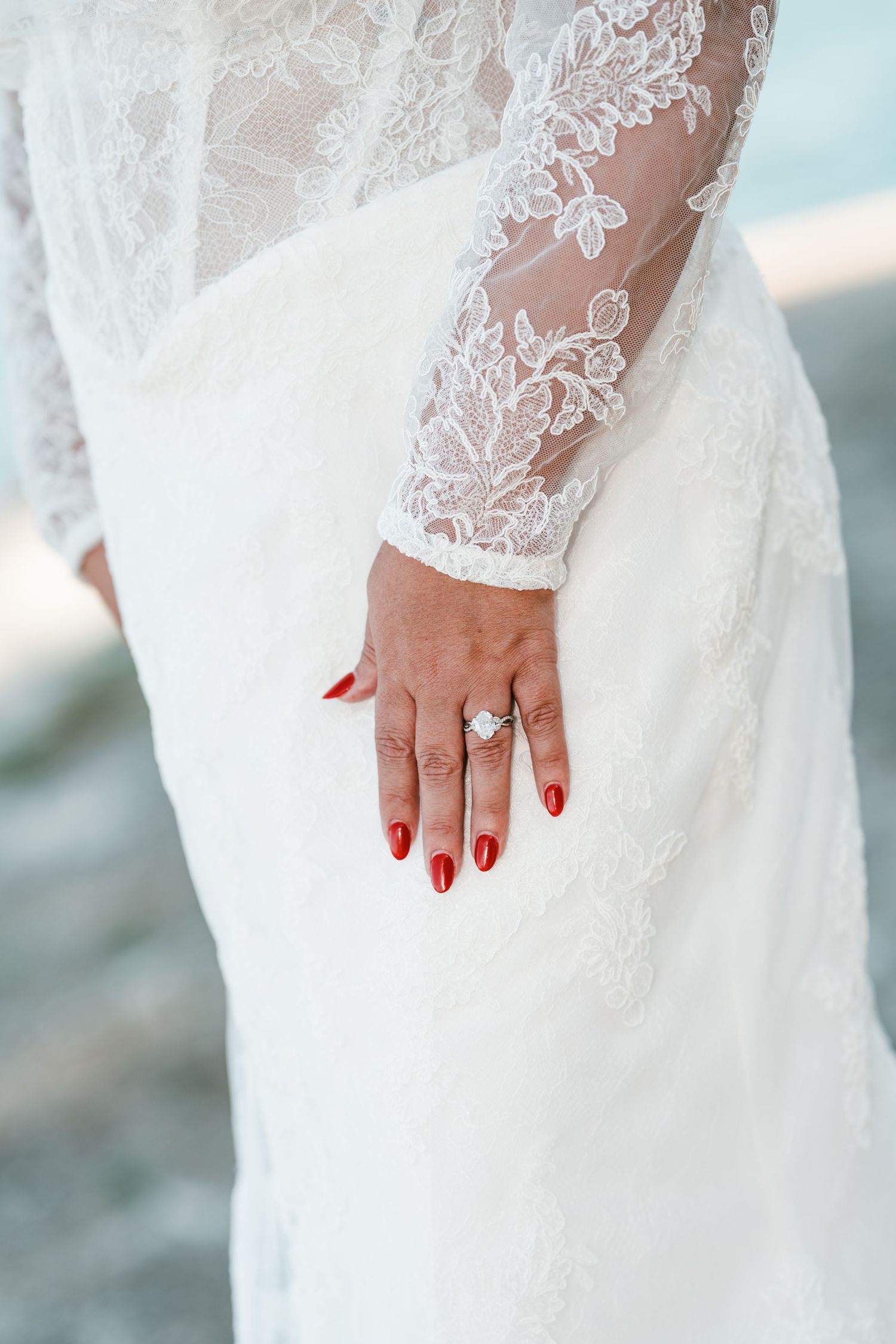 Bride's hand with red nail polish and a diamond engagement ring resting on a white lace wedding dress with sheer long sleeves.