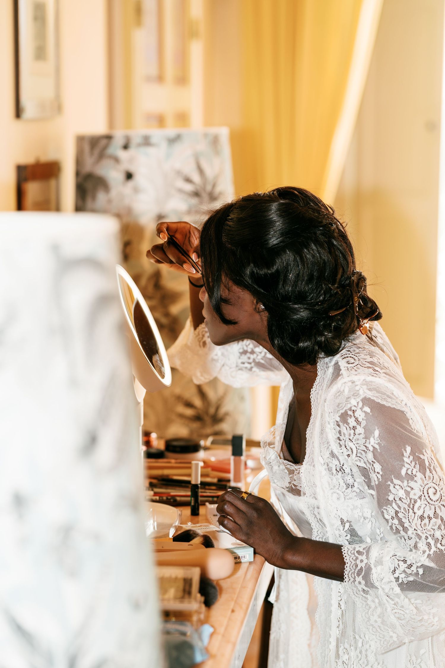 Bride in a white lace robe applies makeup in a small round mirror at a vanity covered with cosmetics, getting ready for her wedding.