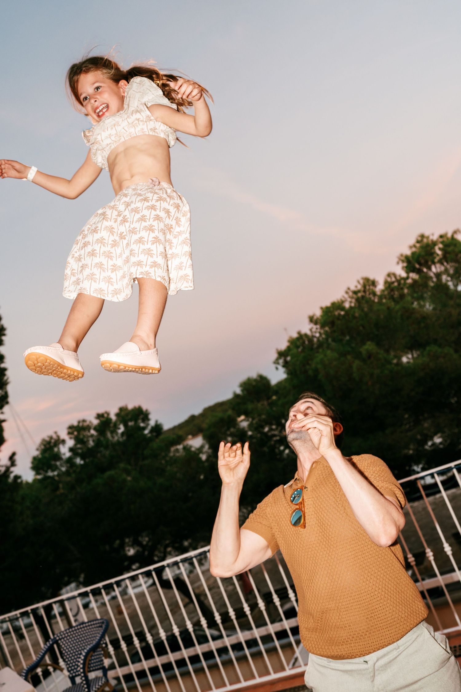Smiling girl in a floral dress is tossed high into the air by a man in an orange polo on a terrace at dusk.