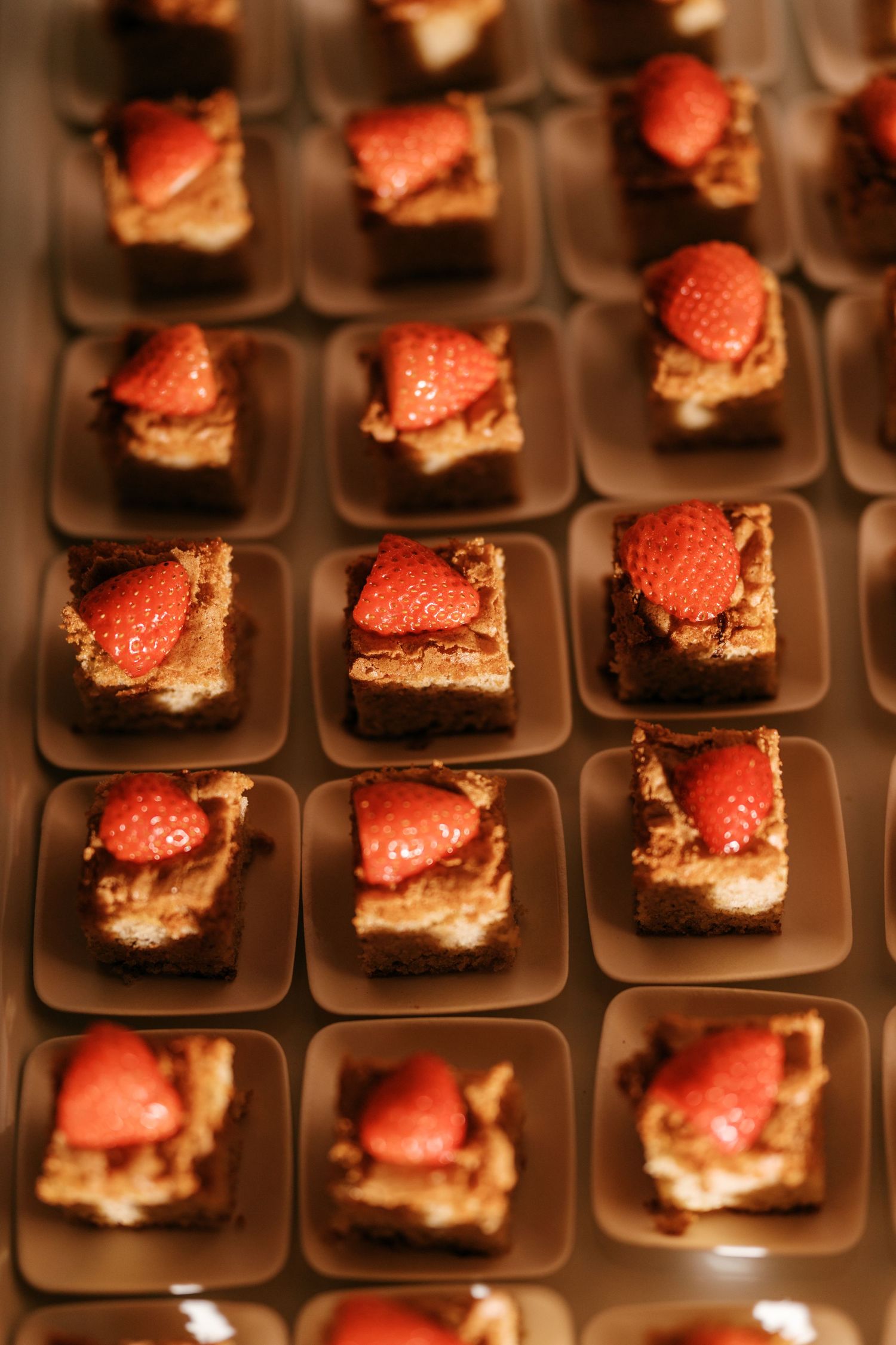 Rows of mini chocolate cake squares topped with fresh strawberries are served on small brown plates at a wedding reception.