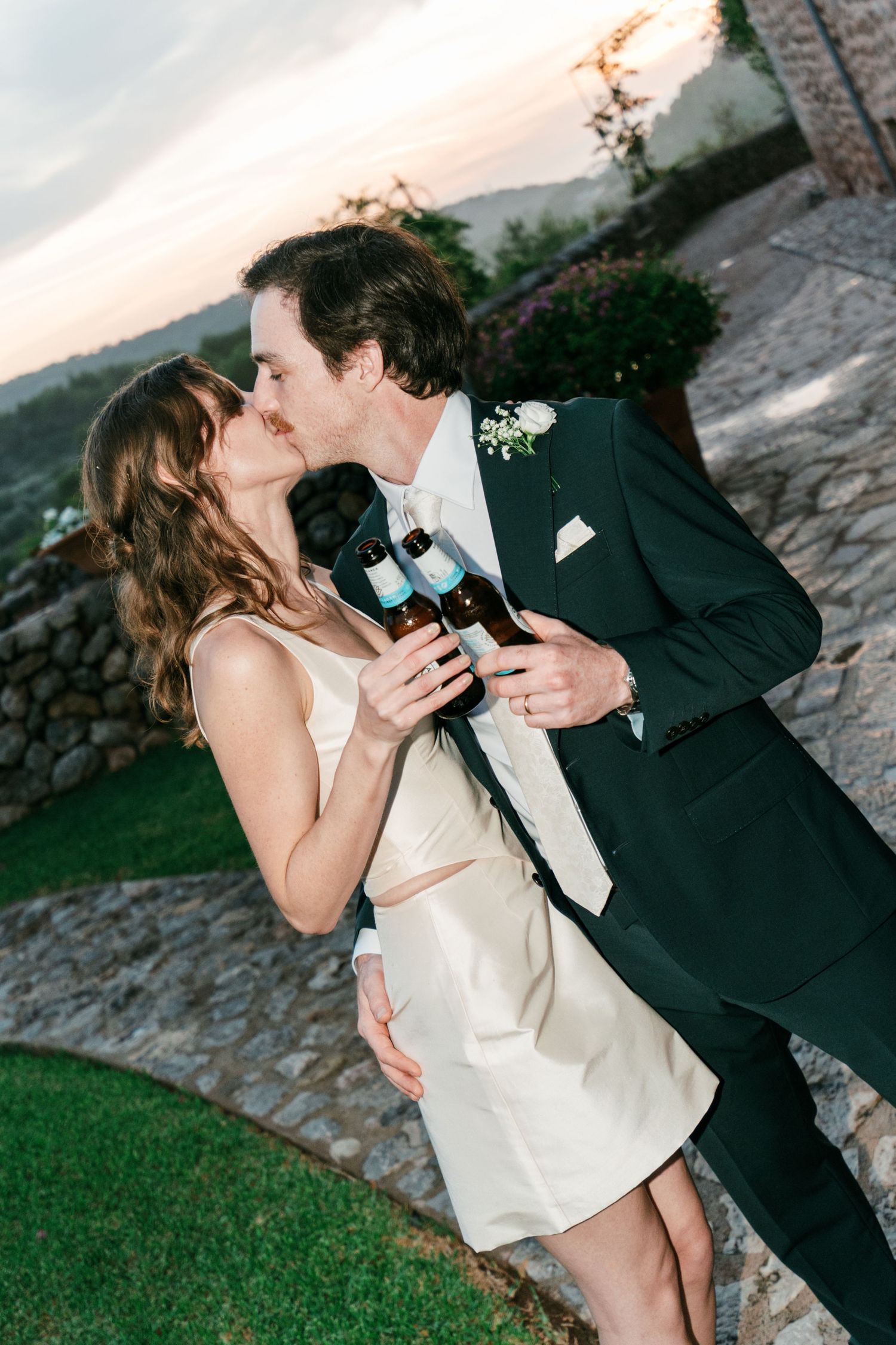 A bride in a cream dress and a groom in a dark suit kiss while holding beer bottles on a stone path at sunset.