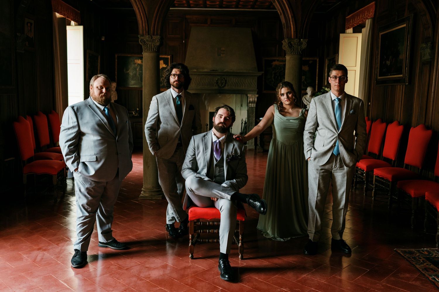Four men in gray suits and a woman in a sage green dress pose in an elegant historic hall with red chairs, one man seated in front.