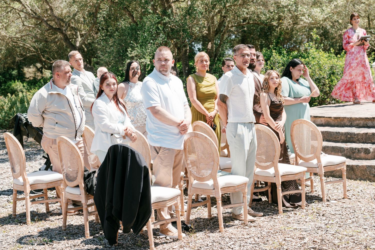 Guests standing outdoors at a wedding ceremony surrounded by lush greenery and elegant wooden chairs in Mallorca.