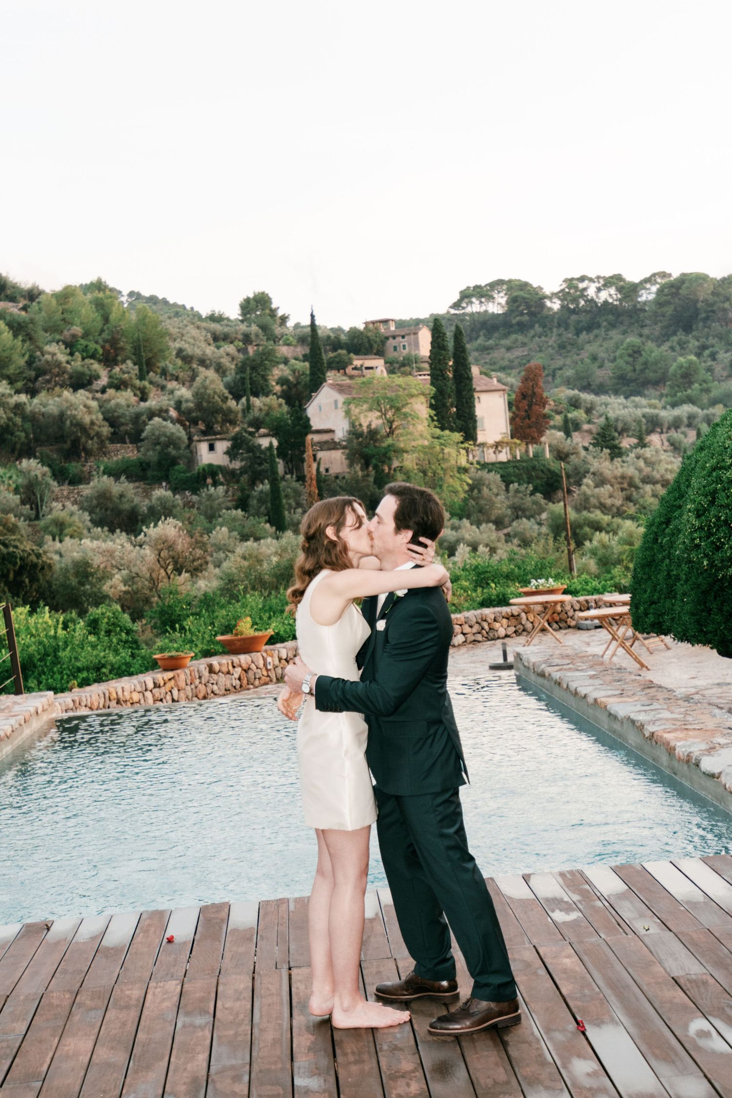 Bride in a short white dress kisses groom in a dark suit on a wooden deck beside a pool with Mediterranean hillside views.