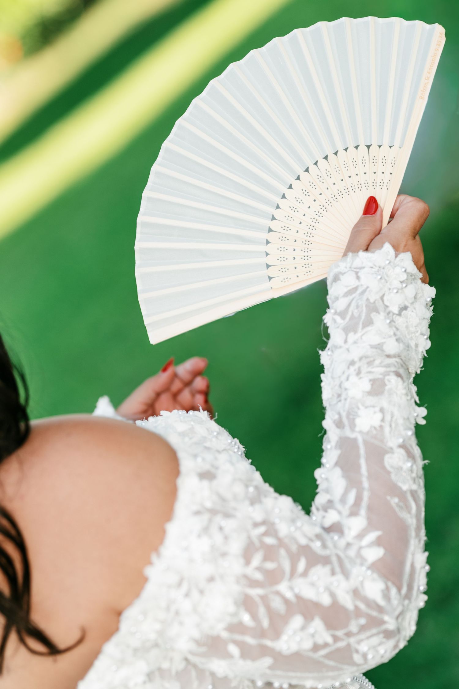 Bride in a white floral lace wedding dress with long sleeves holds up an elegant white folding hand fan against a green background.