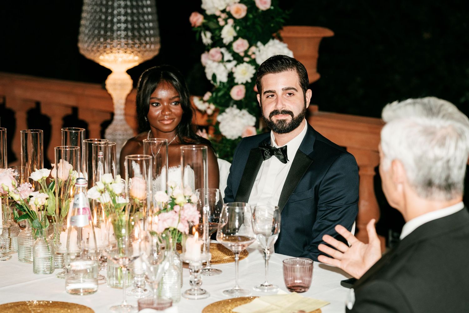 Smiling bride and bearded groom in a tuxedo sit at an evening wedding dinner table with candles, flowers and a guest in foreground.