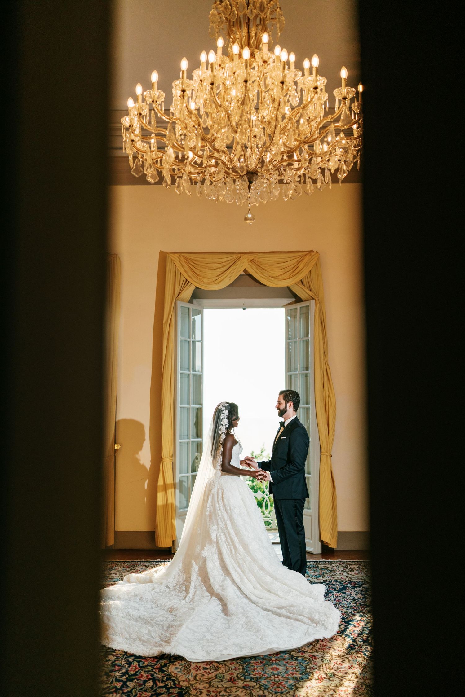 Bride in a lace gown with a long veil and groom in a dark suit exchange vows beneath a grand crystal chandelier in an elegant ballroom.