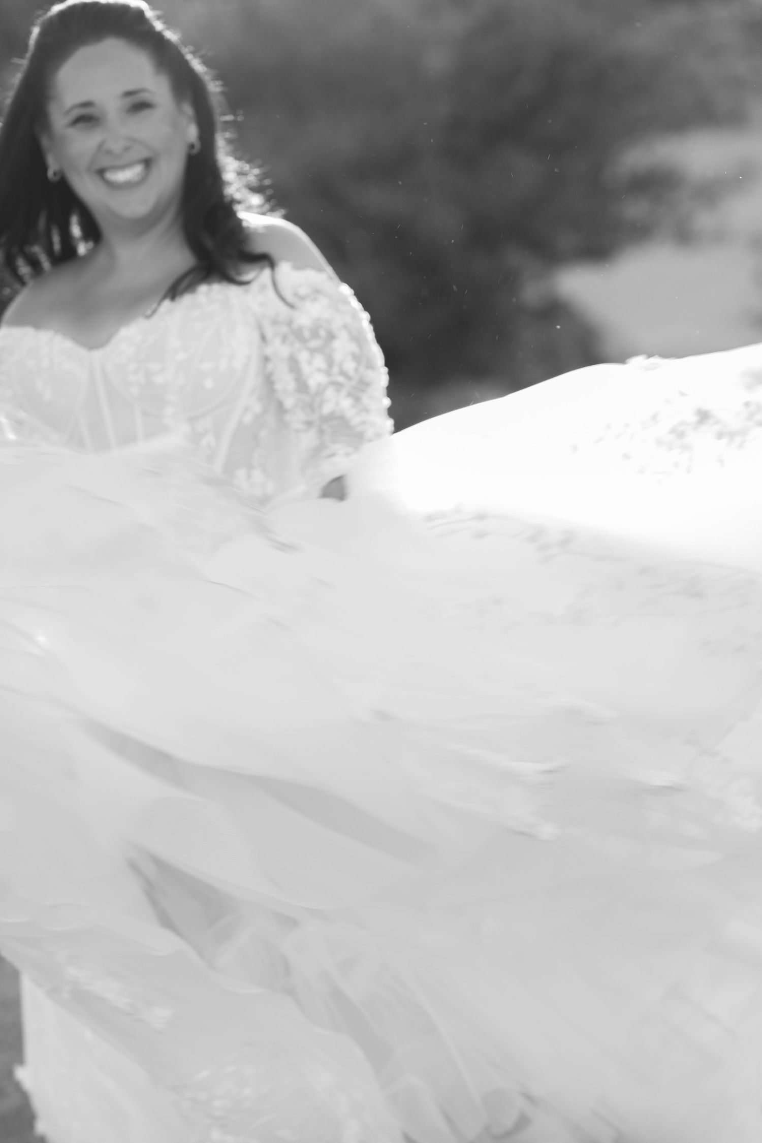Smiling bride in a black and white photo wears a strapless lace wedding gown with a flowing tulle skirt billowing outdoors.