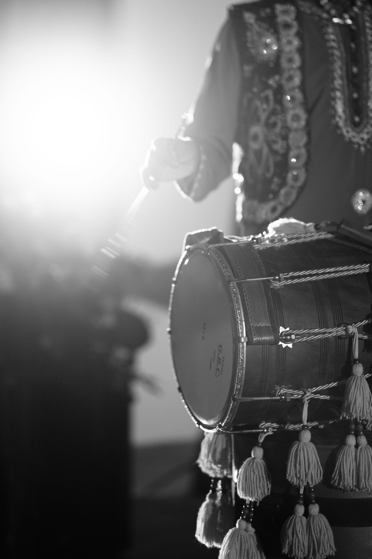 Black and white photo of a dhol drummer in an embroidered jacket playing a tasseled drum with a stick as bright sunlight streams behind.
