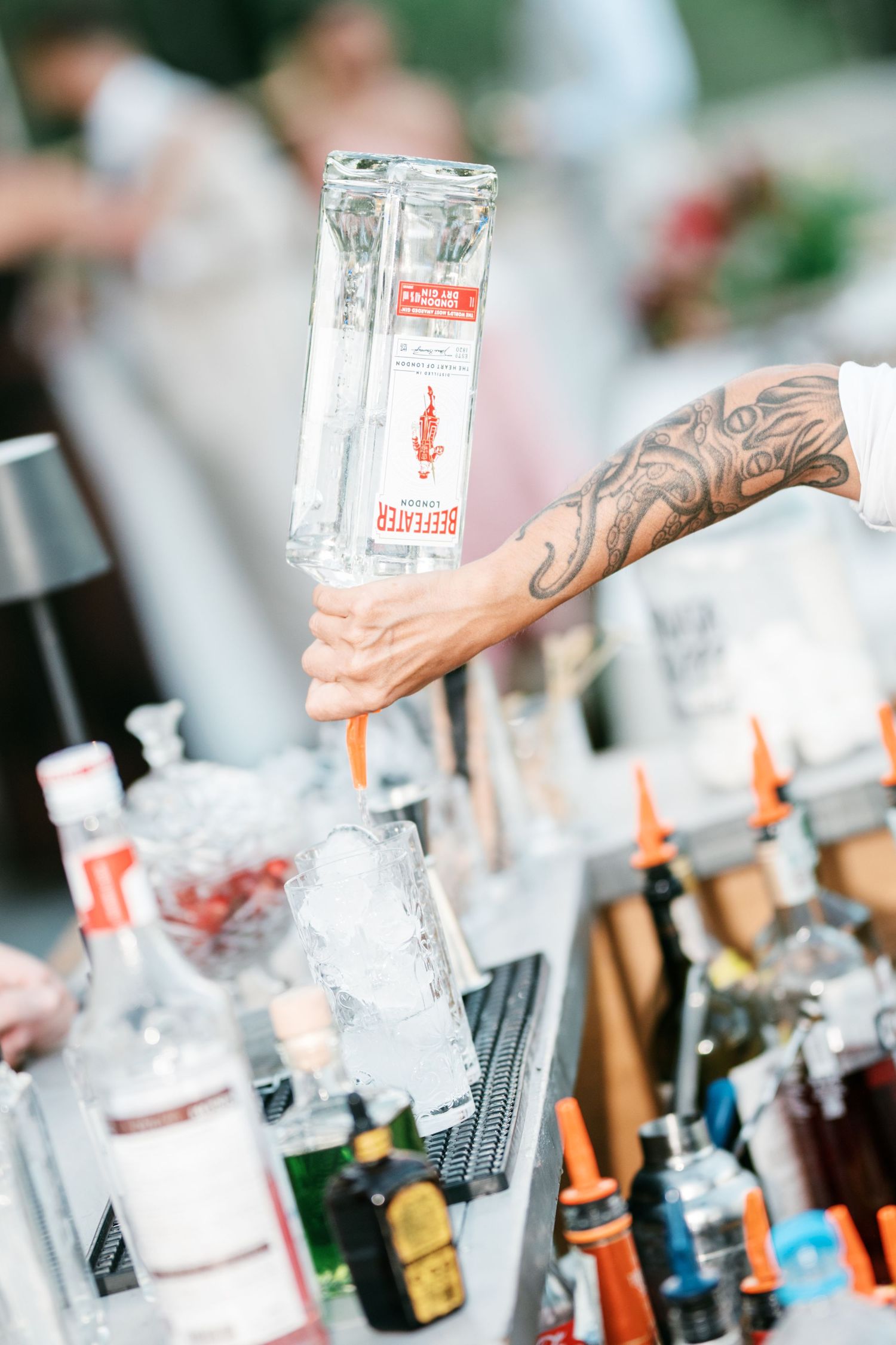 Tattooed bartender pours Beefeater gin from an upside-down bottle into an ice-filled glass at an outdoor event bar.