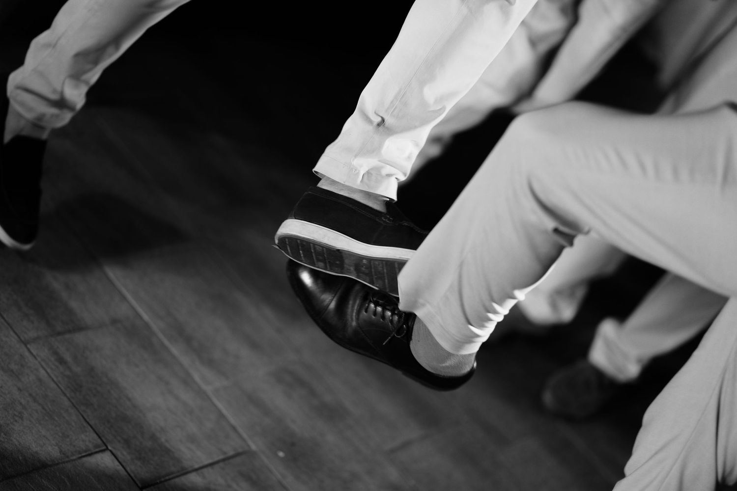 Black and white photo of two people sitting with legs crossed, one foot raised to show a leather dress shoe and light trousers.