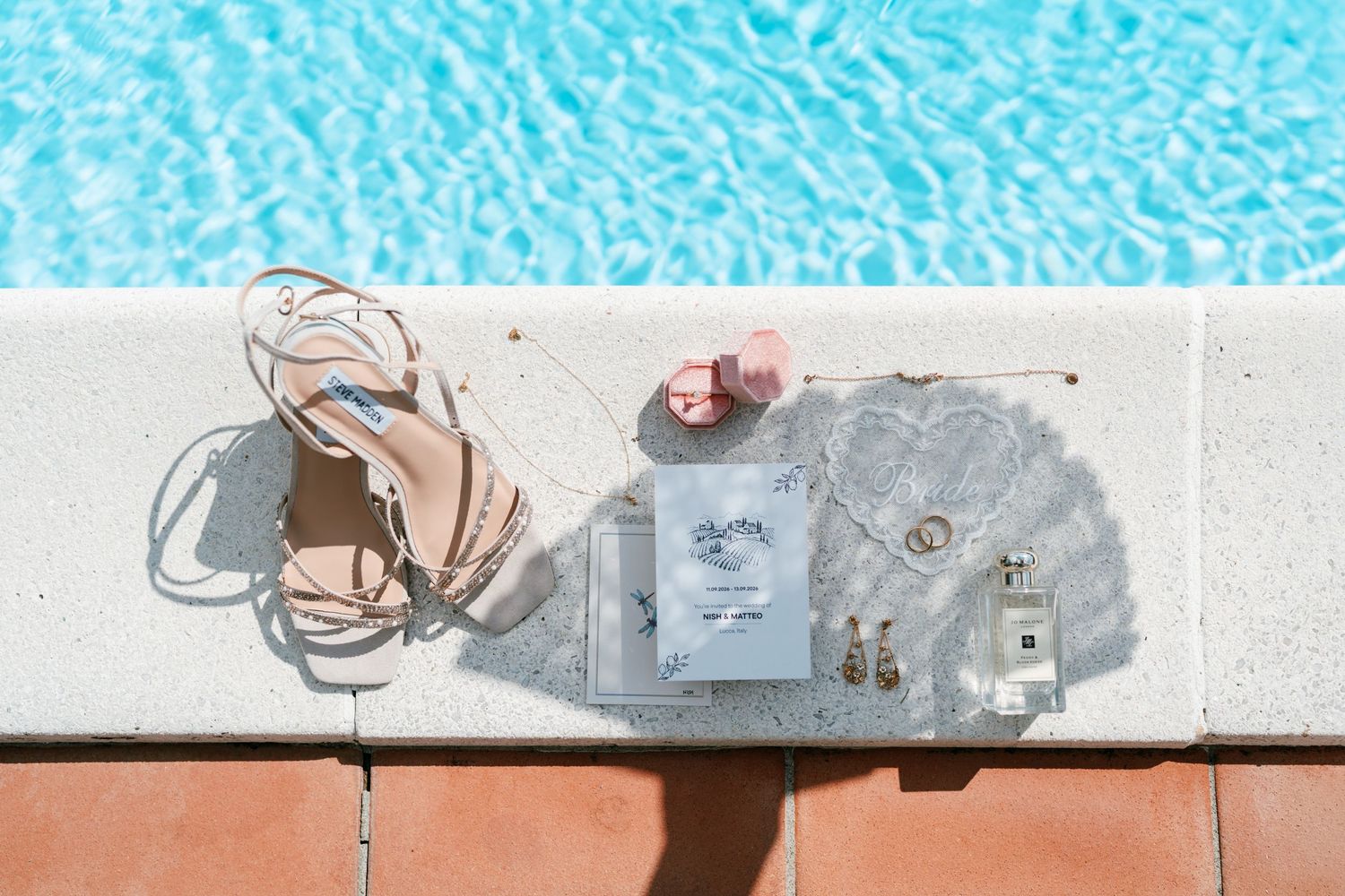 Strappy nude sandals, perfume bottle, earrings and a card styled on a white pool ledge beside sparkling turquoise water.
