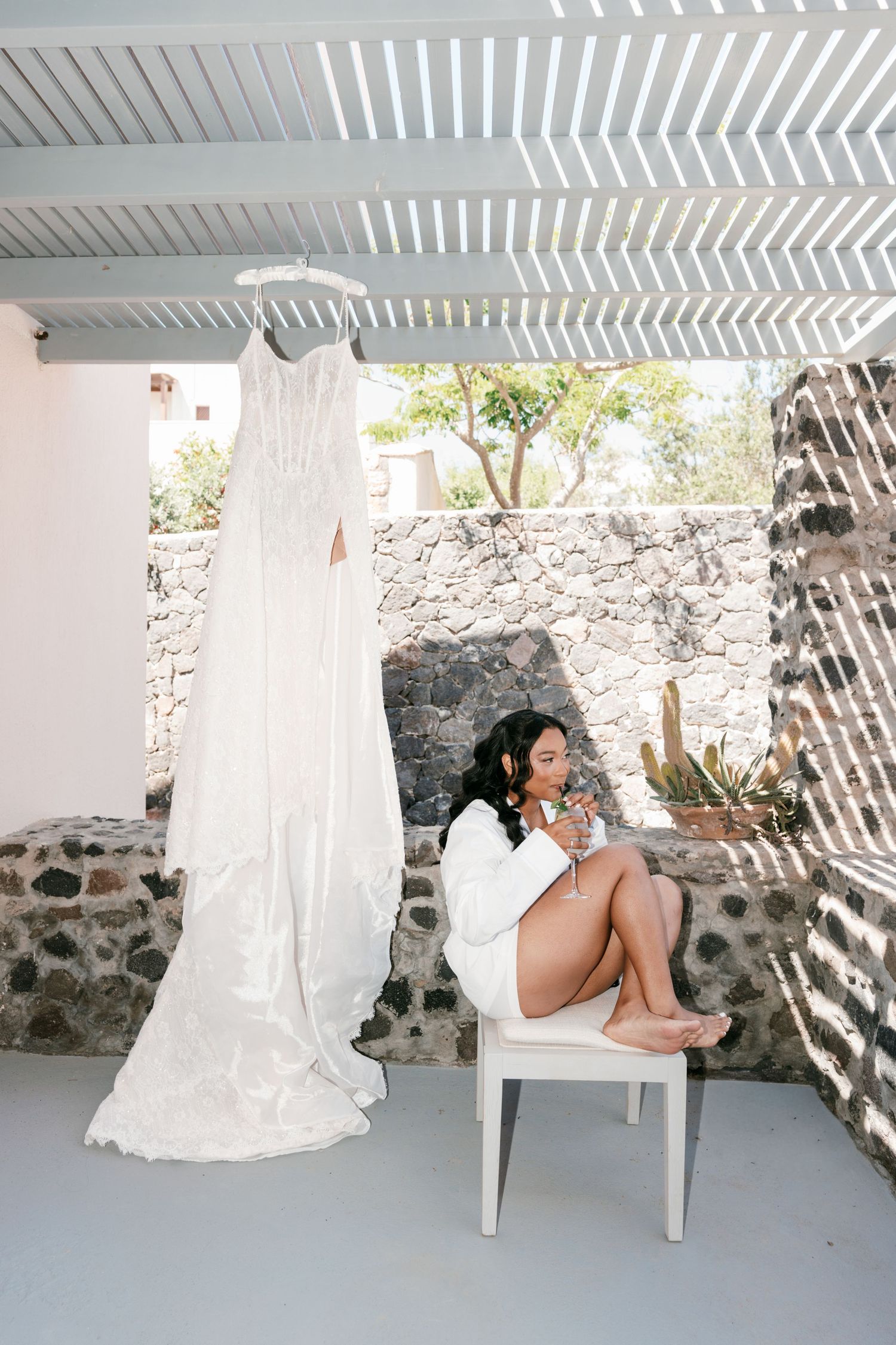 Bride in white robe sits beside her elegant wedding gown hanging outdoors at a Mediterranean venue.