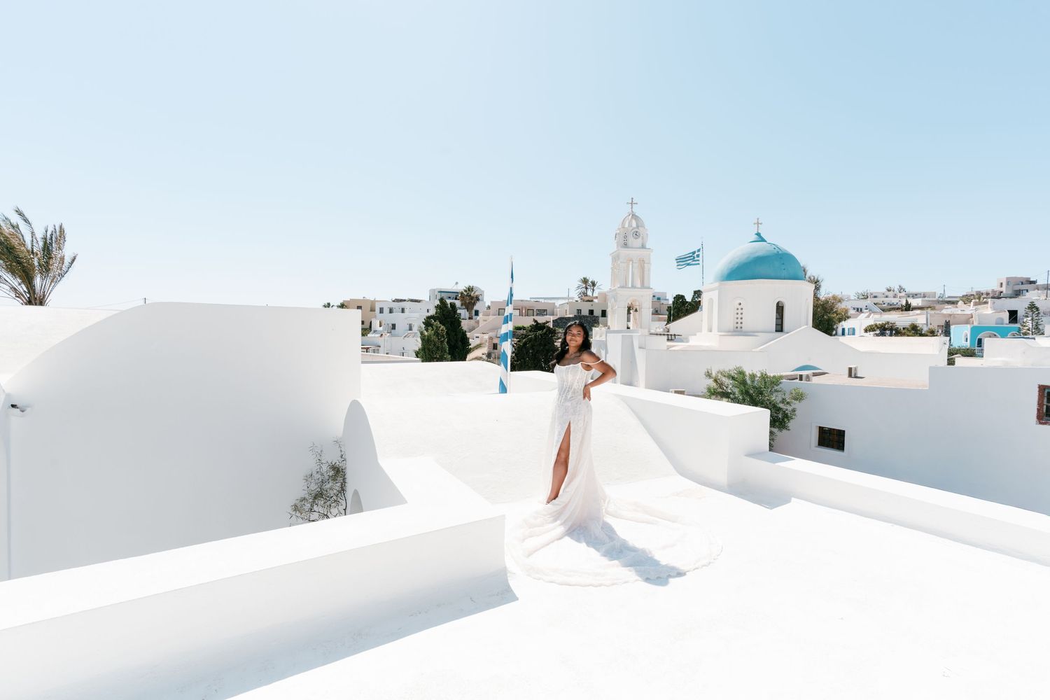 Woman in white dress poses on rooftop in Santorini Greece with blue-domed church and whitewashed buildings in background.