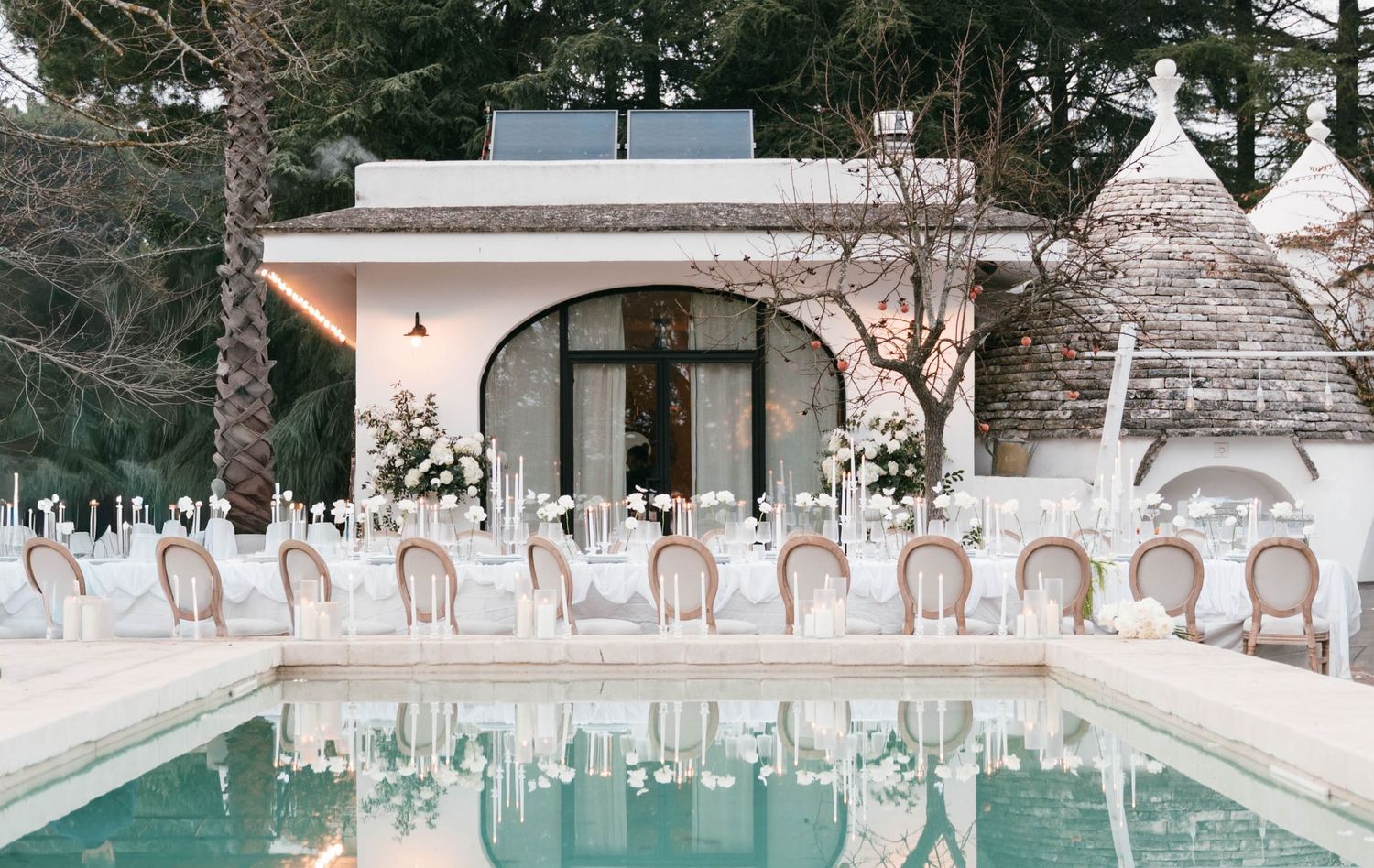 Elegant outdoor wedding reception setup beside a pool with white chairs and floral centerpieces near a trullo villa.Puglia wedding photograp