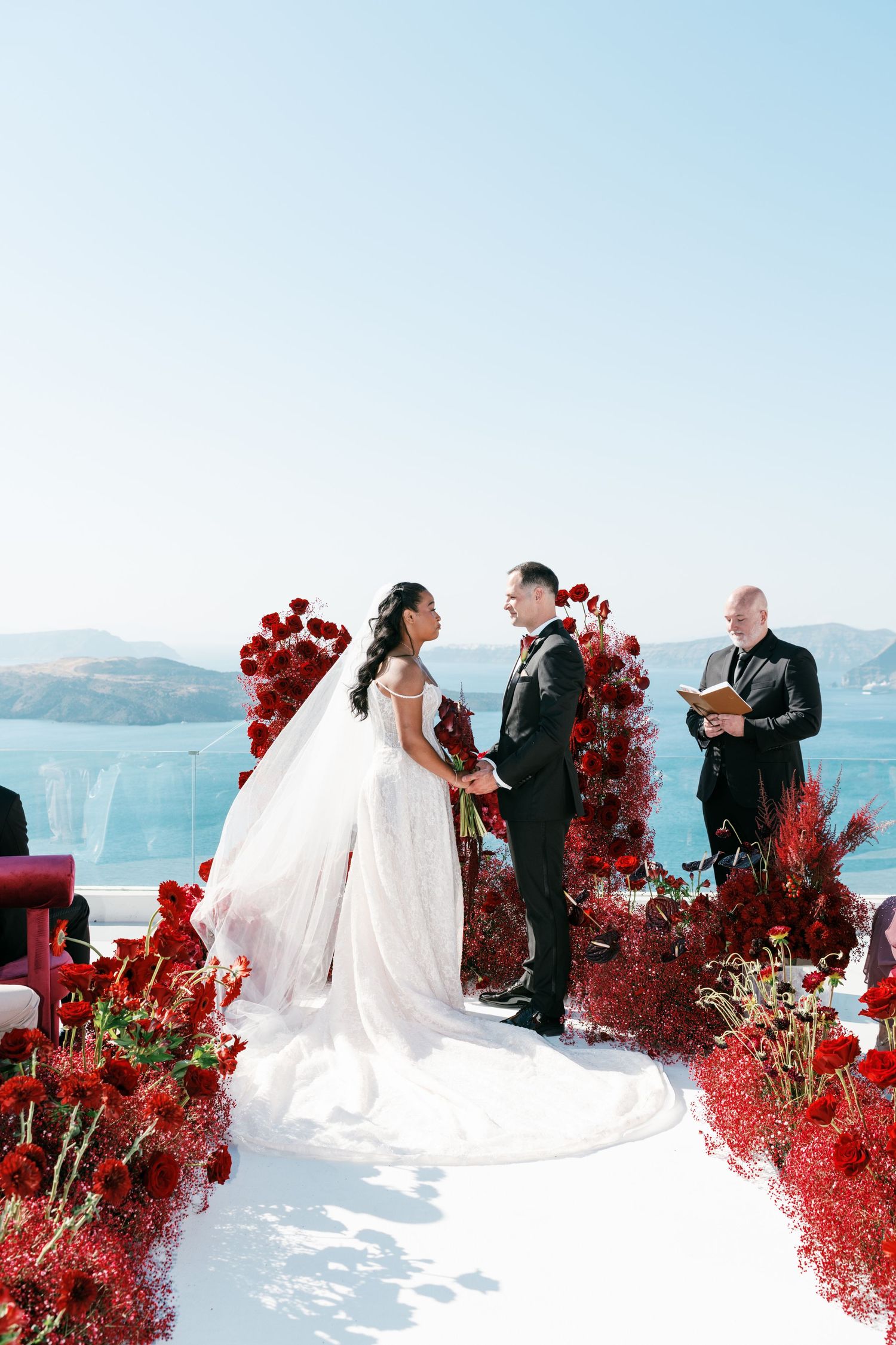Bride and groom exchange vows at an outdoor wedding ceremony adorned with red roses, overlooking a stunning ocean view.