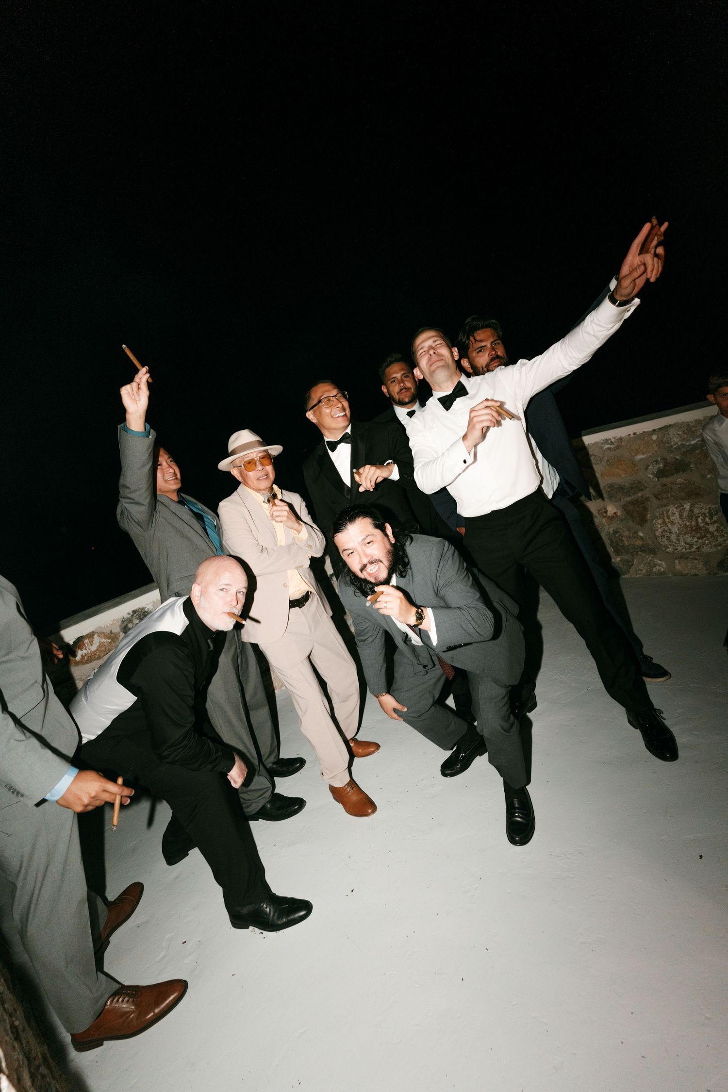 Group of men in formal attire celebrating at a nighttime wedding reception, posing energetically outdoors.