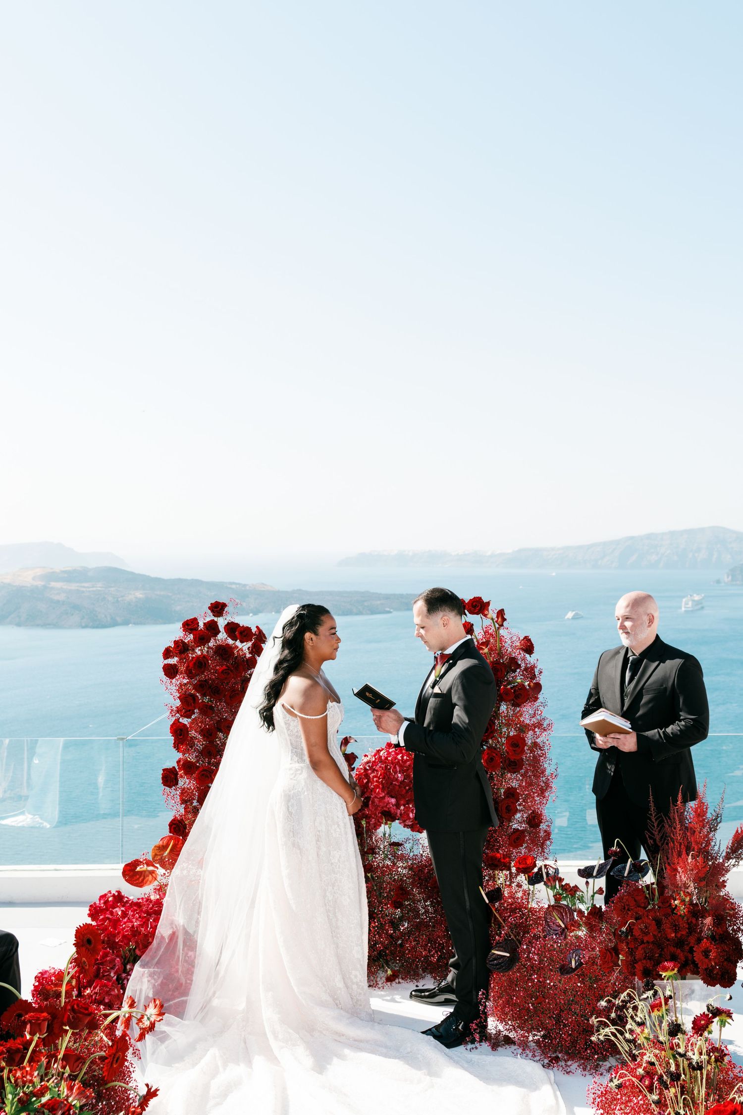 Bride and groom exchange vows at a stunning Santorini wedding ceremony with red floral arch and caldera sea views.