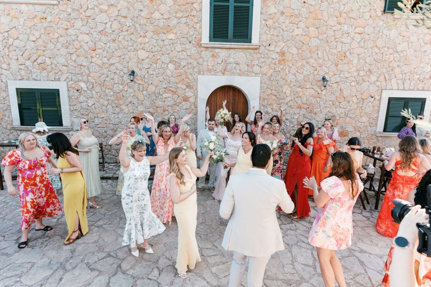 Wedding guests celebrate outdoors at a rustic stone venue, with the bride and groom surrounded by colorfully dressed attendees. Mallorca