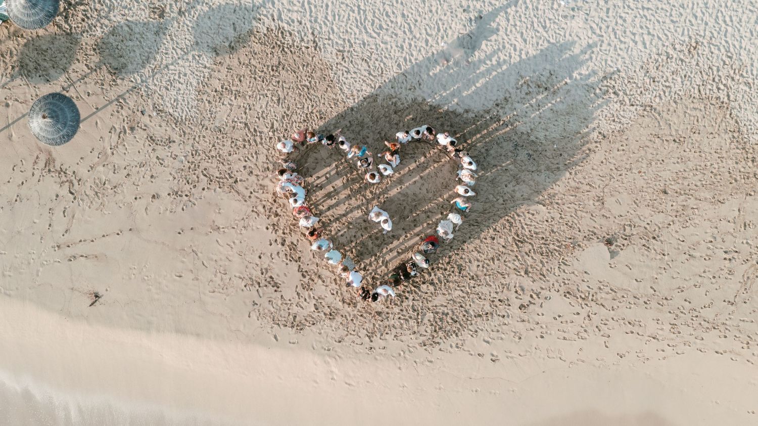 Aerial view of people forming a heart shape on a sandy beach near the ocean shoreline. Mallorca wedding filmmaker, videographer