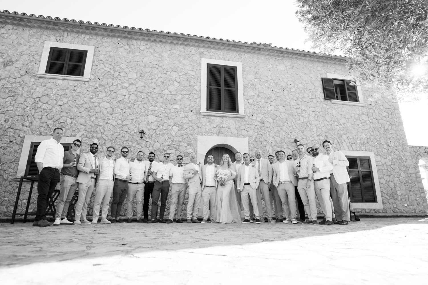 Black and white wedding photo of large bridal party posing in front of rustic stone building. Mallorca wedding photographer