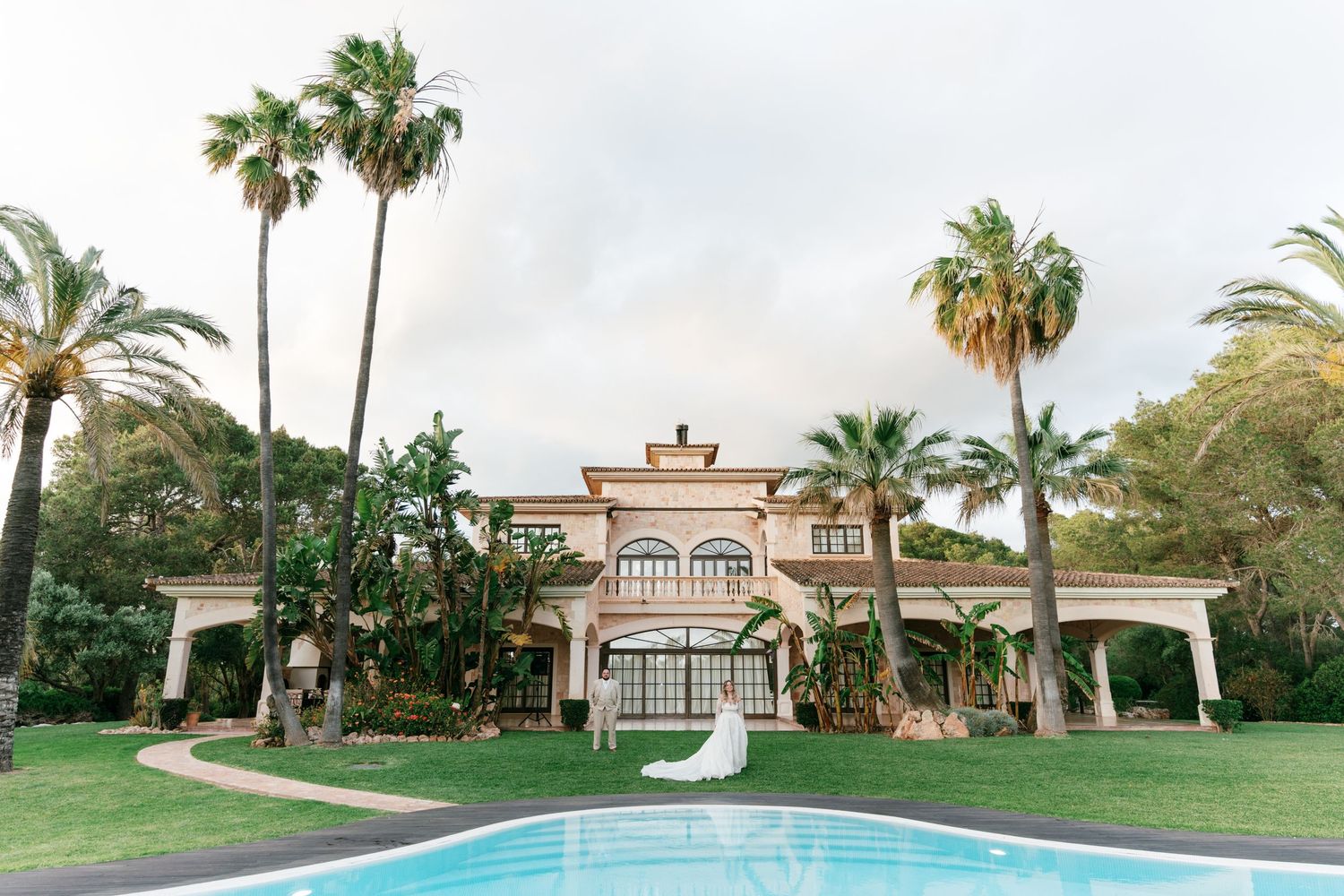 Bride poses in front of elegant Mediterranean villa with palm trees and pool in lush garden setting in Mallorca.