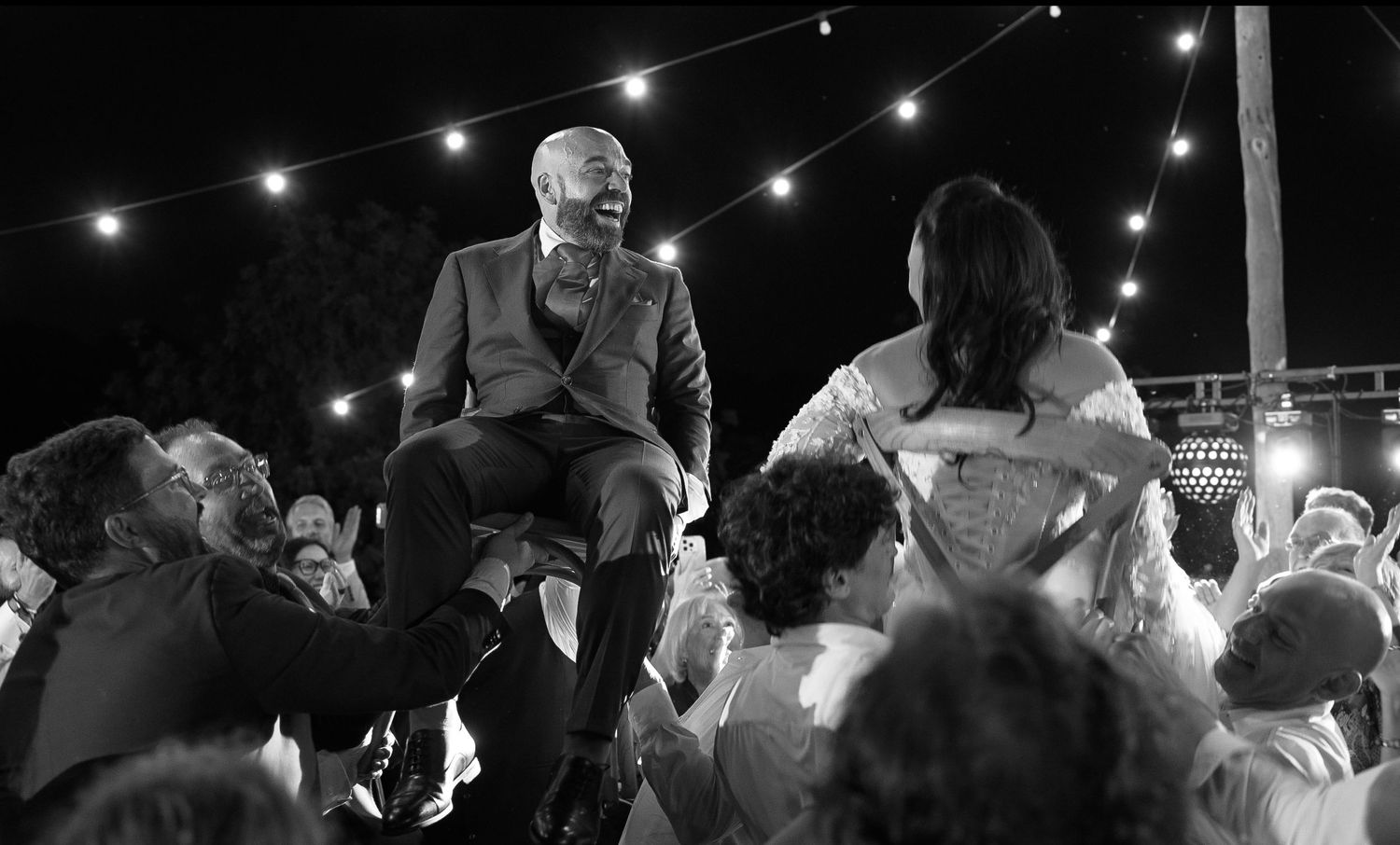 Black and white photo of a smiling groom and bride lifted on chairs by cheering guests at a lively wedding reception under string lights.
