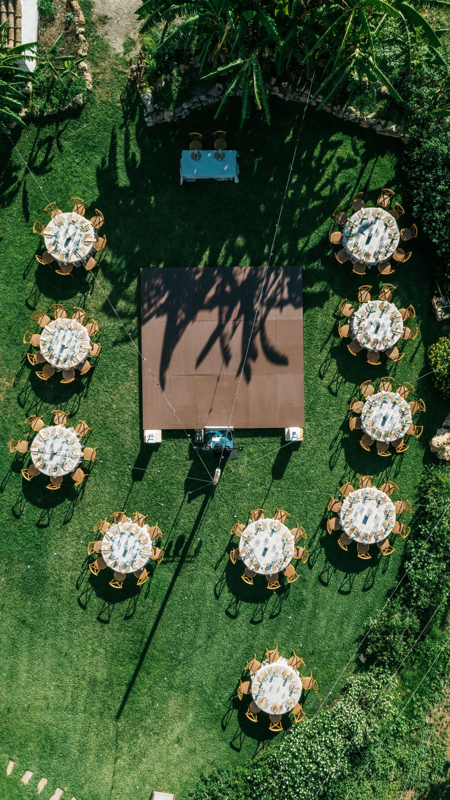 Aerial view of an outdoor wedding reception on a green lawn with round tables, wooden chairs, a central dance floor and a head table.