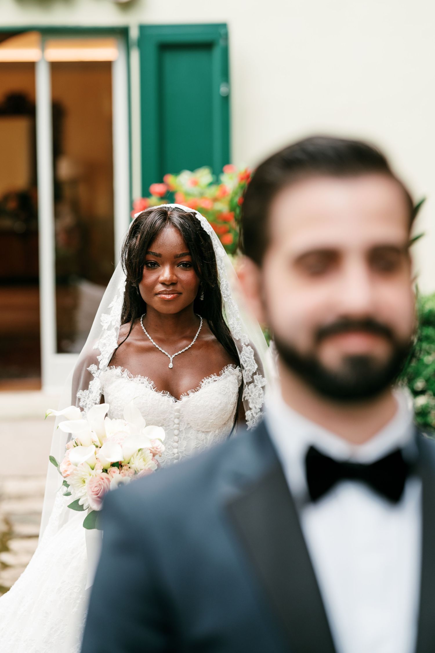 Bride in a lace gown and veil holds a white and pink bouquet as a blurred groom in a tuxedo and bow tie stands in the foreground.