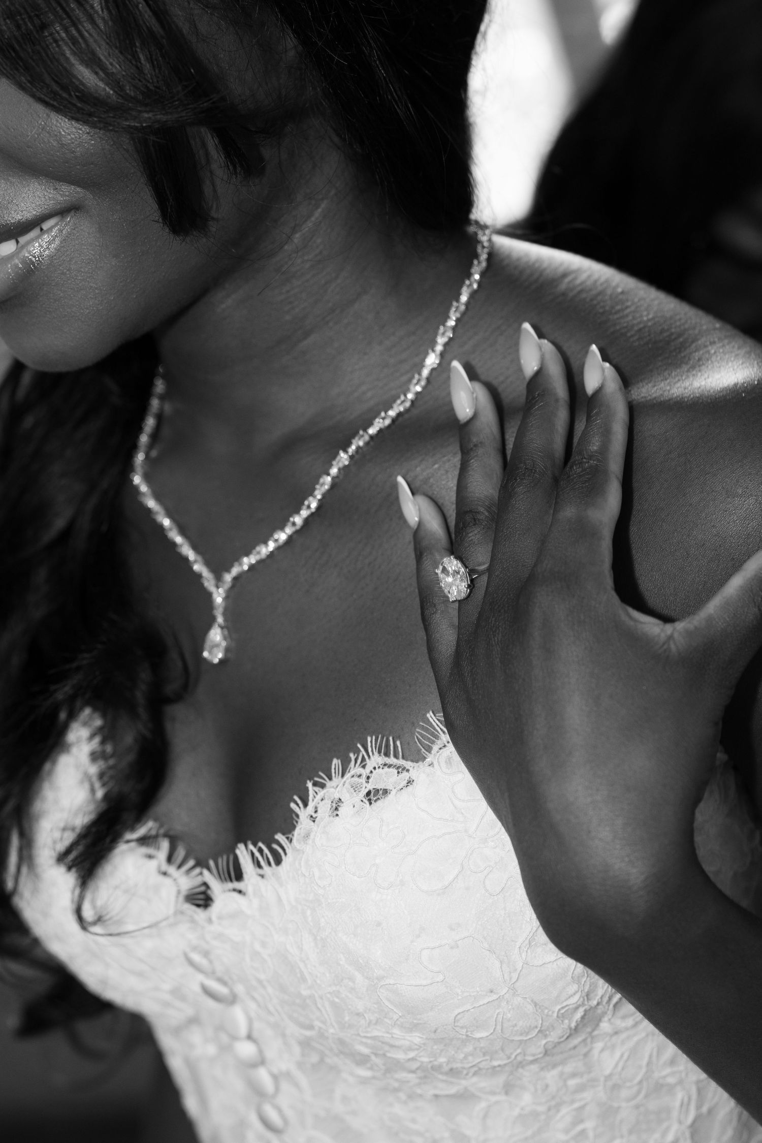 Black and white close-up of a bride in a lace wedding gown showing her diamond ring and wearing a sparkling pendant necklace.