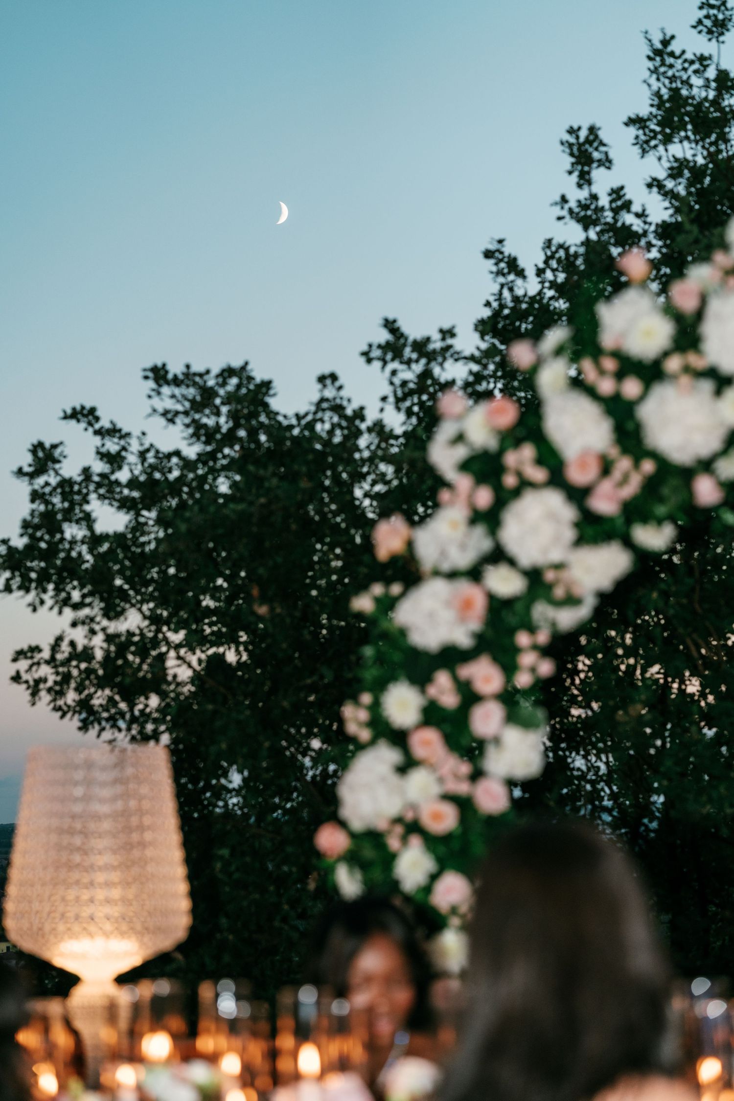 Blurred wedding guests beneath a crescent moon at dusk, with a floral arch of white and blush roses and a glowing lamp beside dark trees.